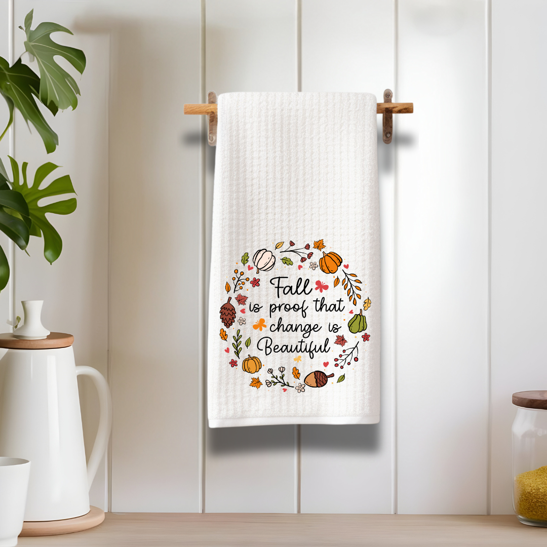 A farmhouse-style kitchen scene with a fall-themed kitchen towel hanging on a wooden rack against a white panel wall, styled with neutral decor and greenery for a warm and inviting aesthetic.