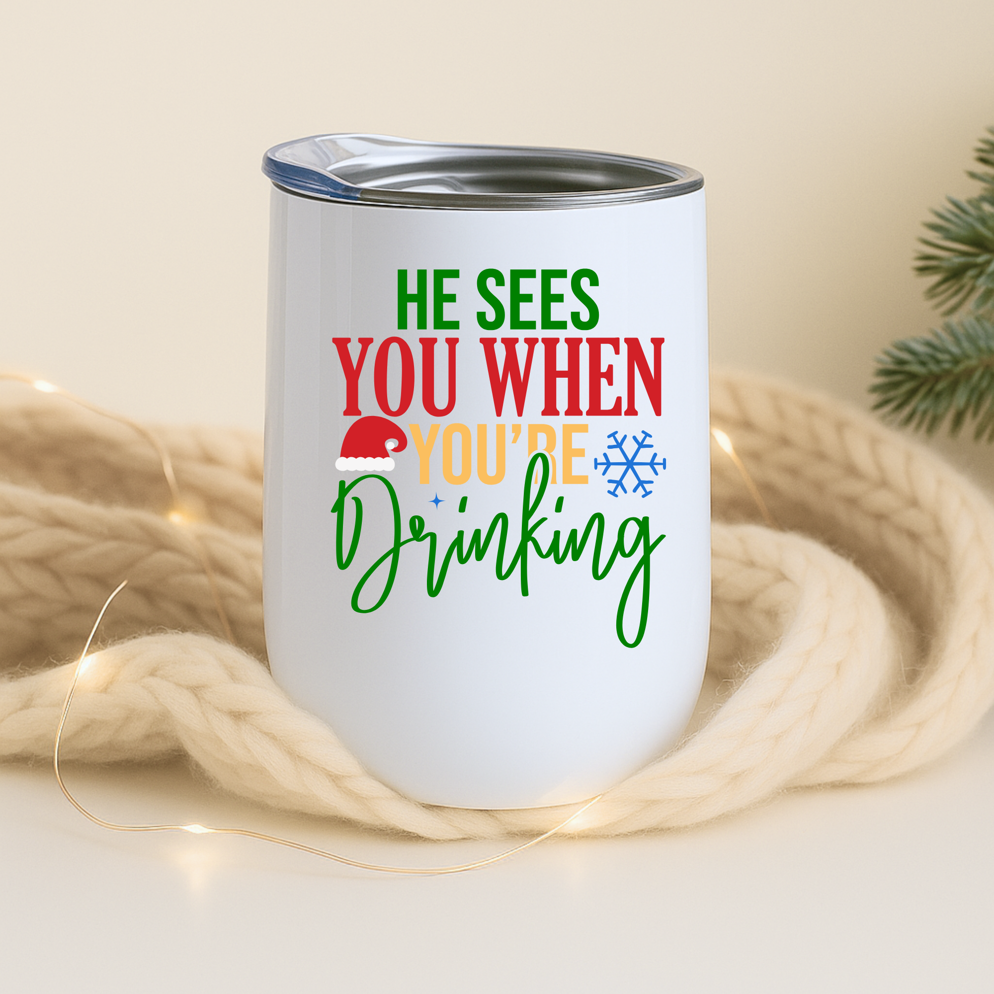 White holiday tumbler with “He Sees You When You’re Drinking” shown on cozy beige knit background with warm string lights.