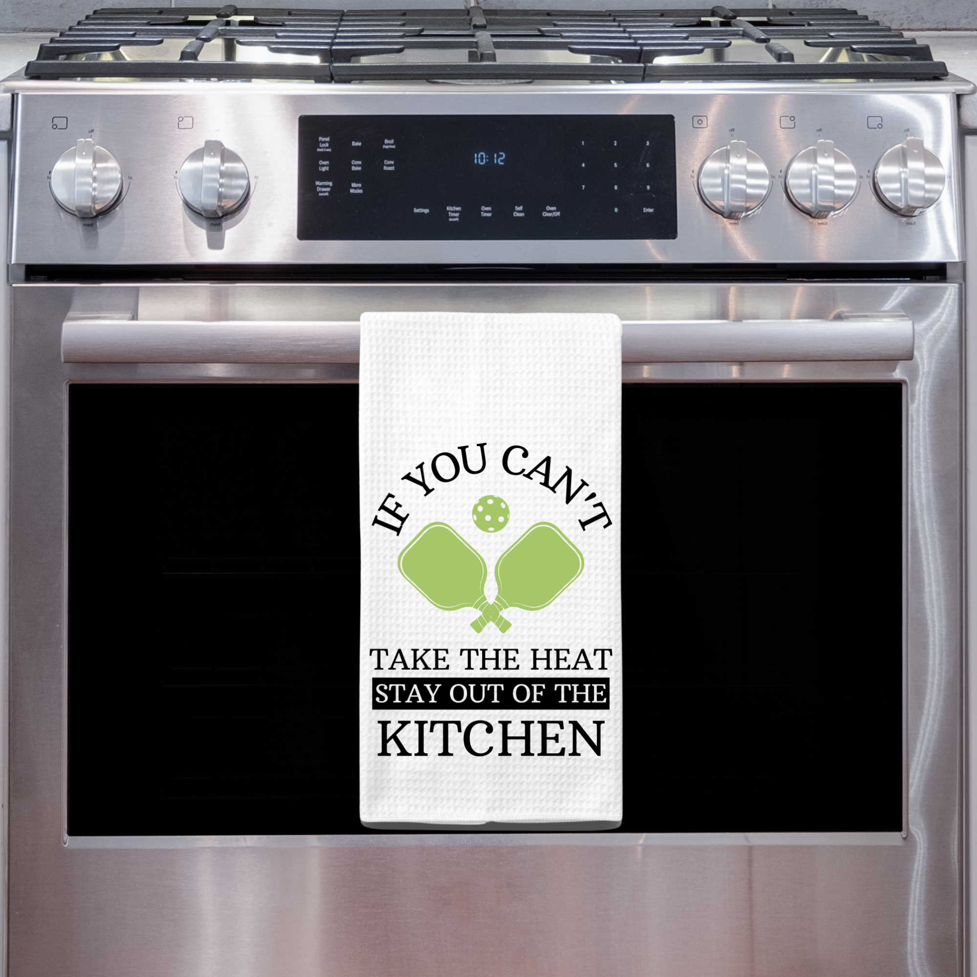 White waffle weave towel with funny pickleball quote hanging on a stainless steel oven in a modern kitchen.