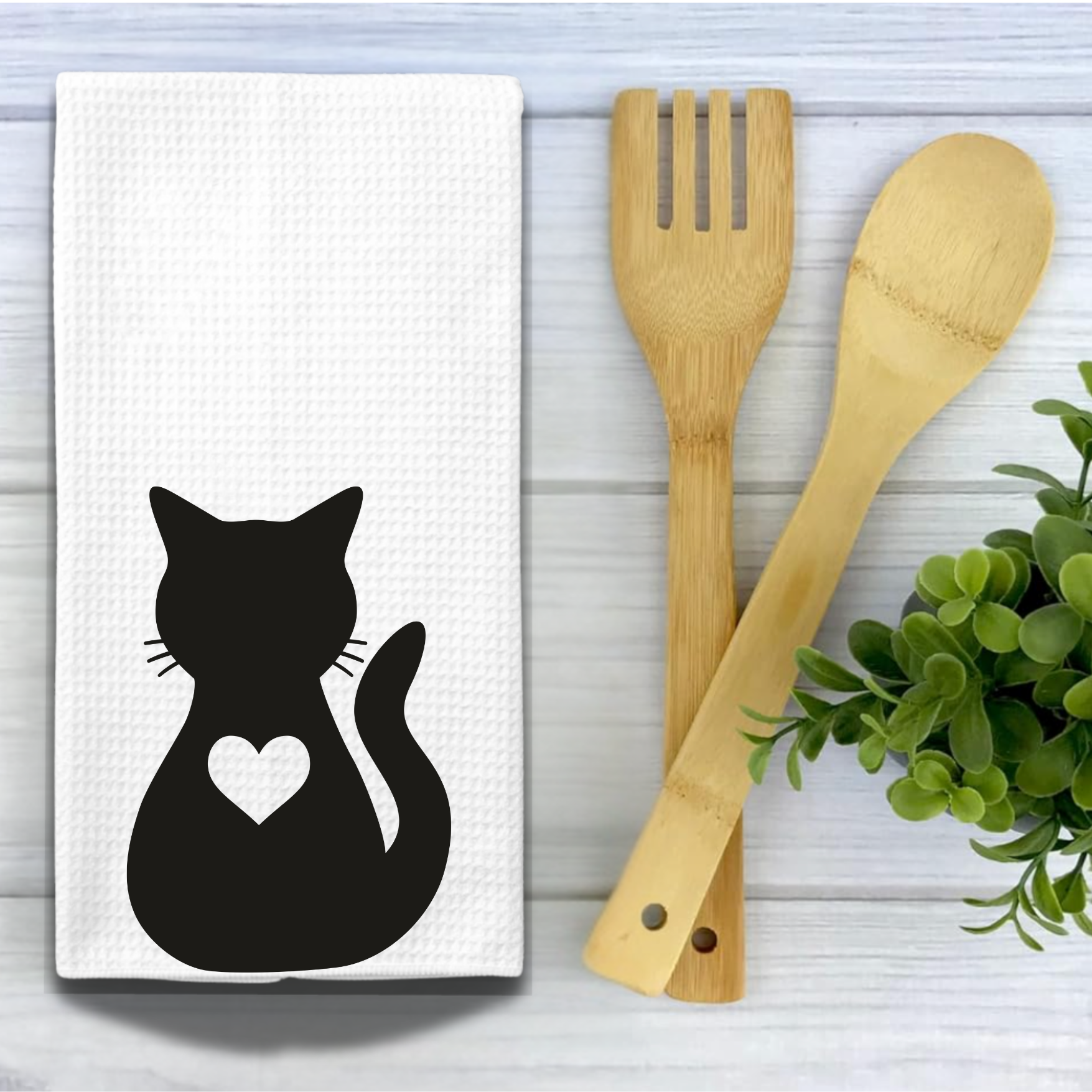 Farmhouse-style tea towel with a black cat design, displayed with bamboo utensils and fresh greenery on a wooden surface.