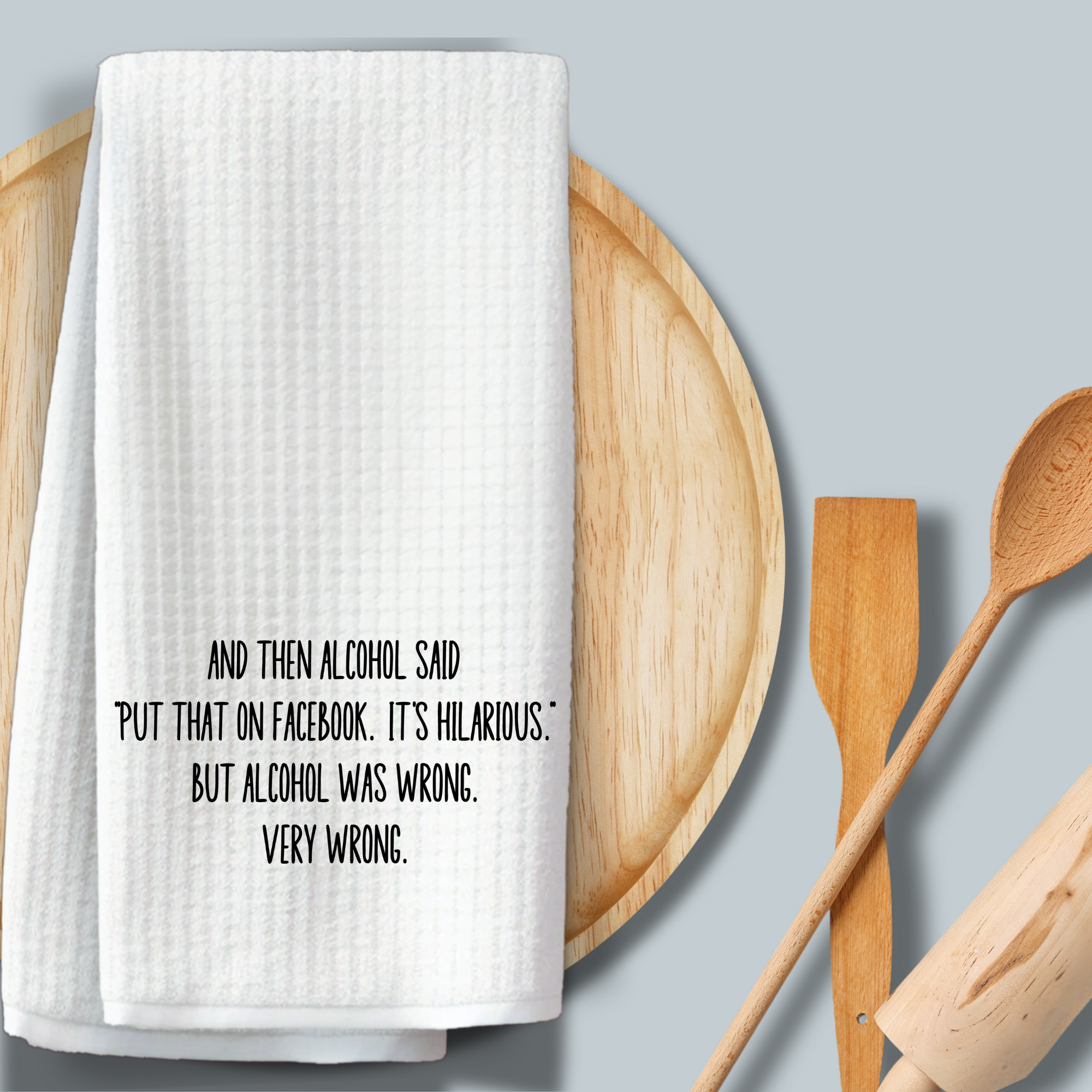 Witty waffle weave kitchen towel with permanent ink design, draped over a wooden tray with cooking utensils.