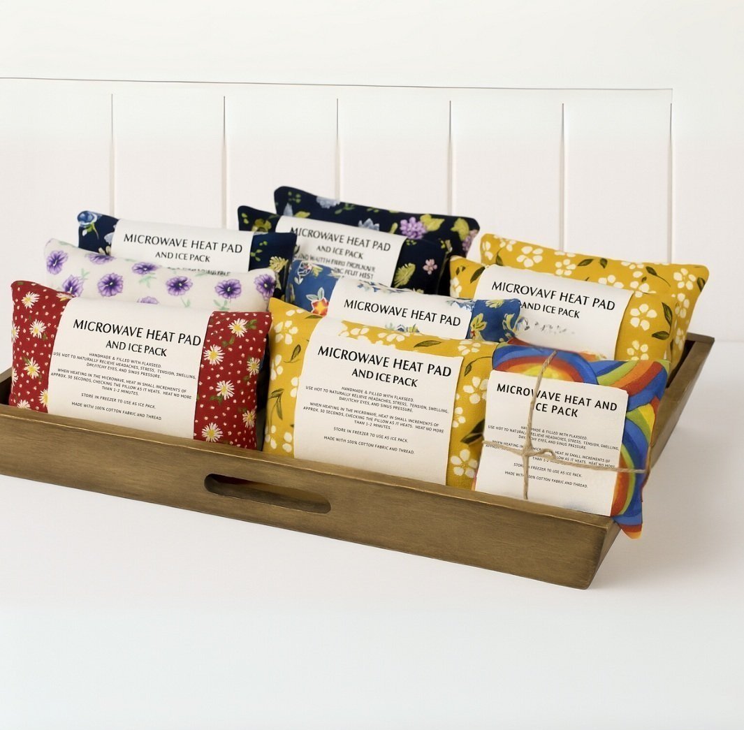 Handmade microwave flaxseed heat pads in colorful floral fabric designs displayed in wooden tray