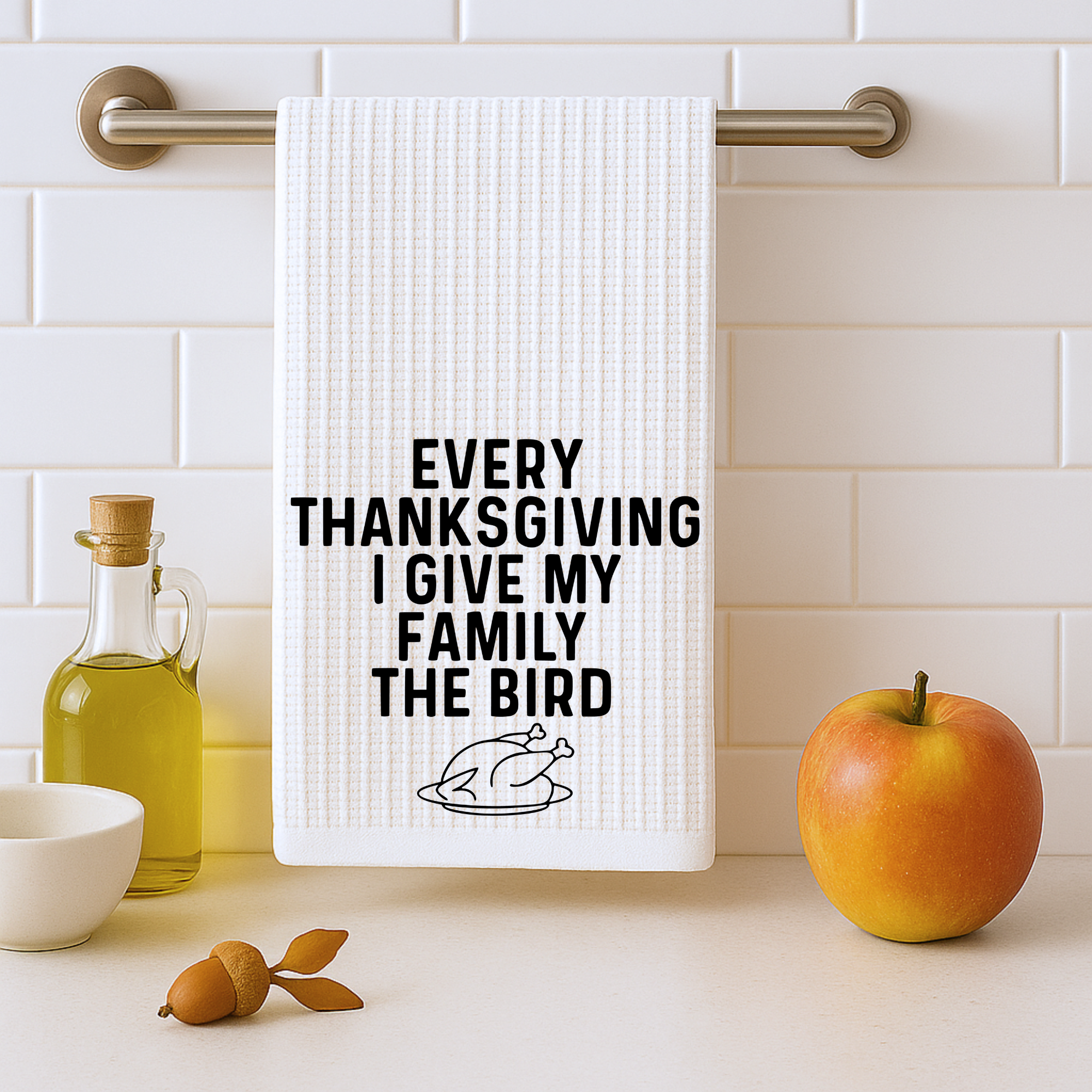 White hand towel hanging on a kitchen bar with apple and olive oil decor, printed with the phrase “Every Thanksgiving I Give My Family the Bird.”