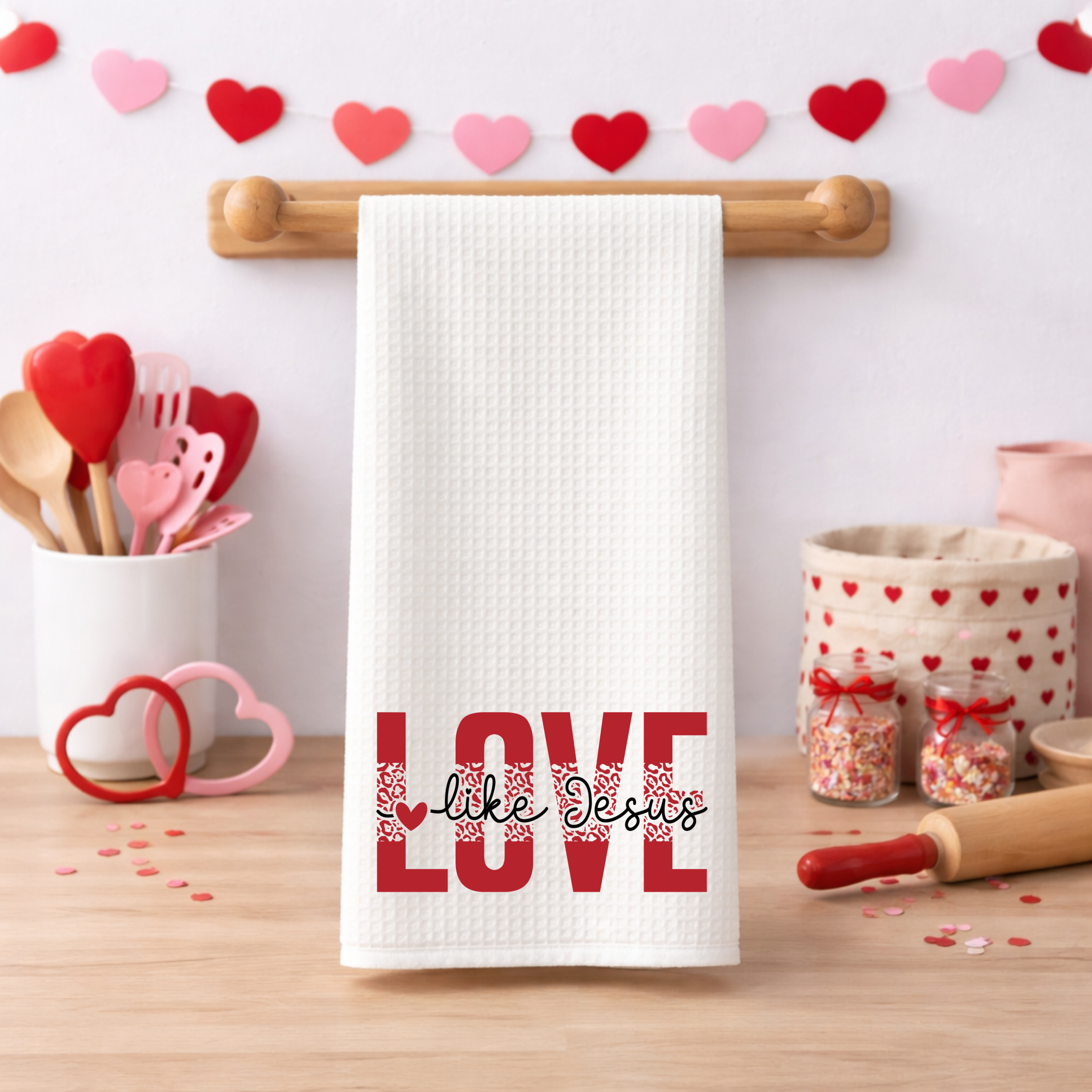 Christian “Love Like Jesus” kitchen towel displayed with heart garland and Valentine baking props.
