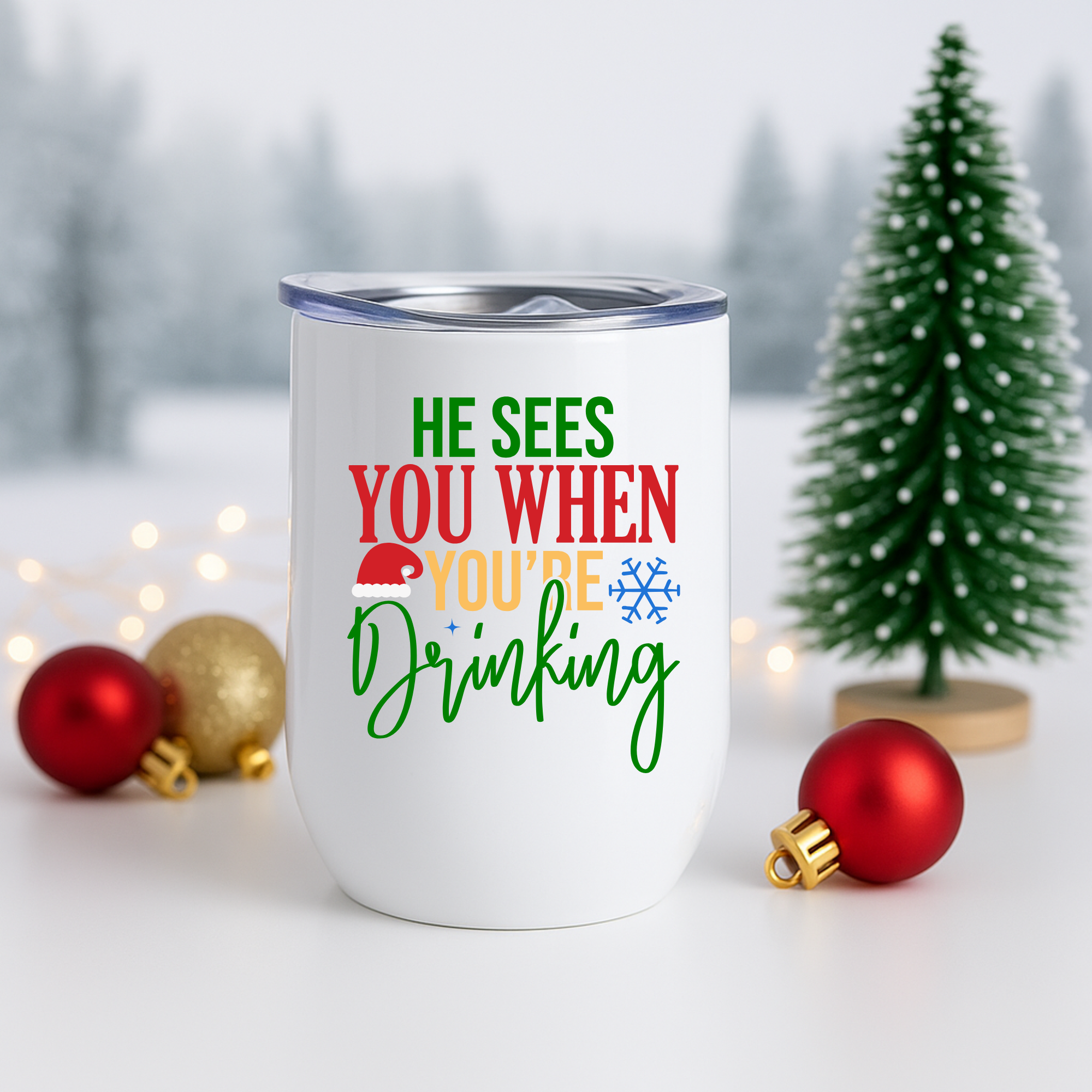 Festive stainless tumbler with “He Sees You When You’re Drinking” displayed beside red and gold ornaments and a Christmas tree.