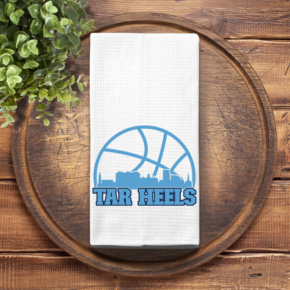 Tar Heels basketball kitchen towel styled on round wood tray with faux plant for rustic presentation.