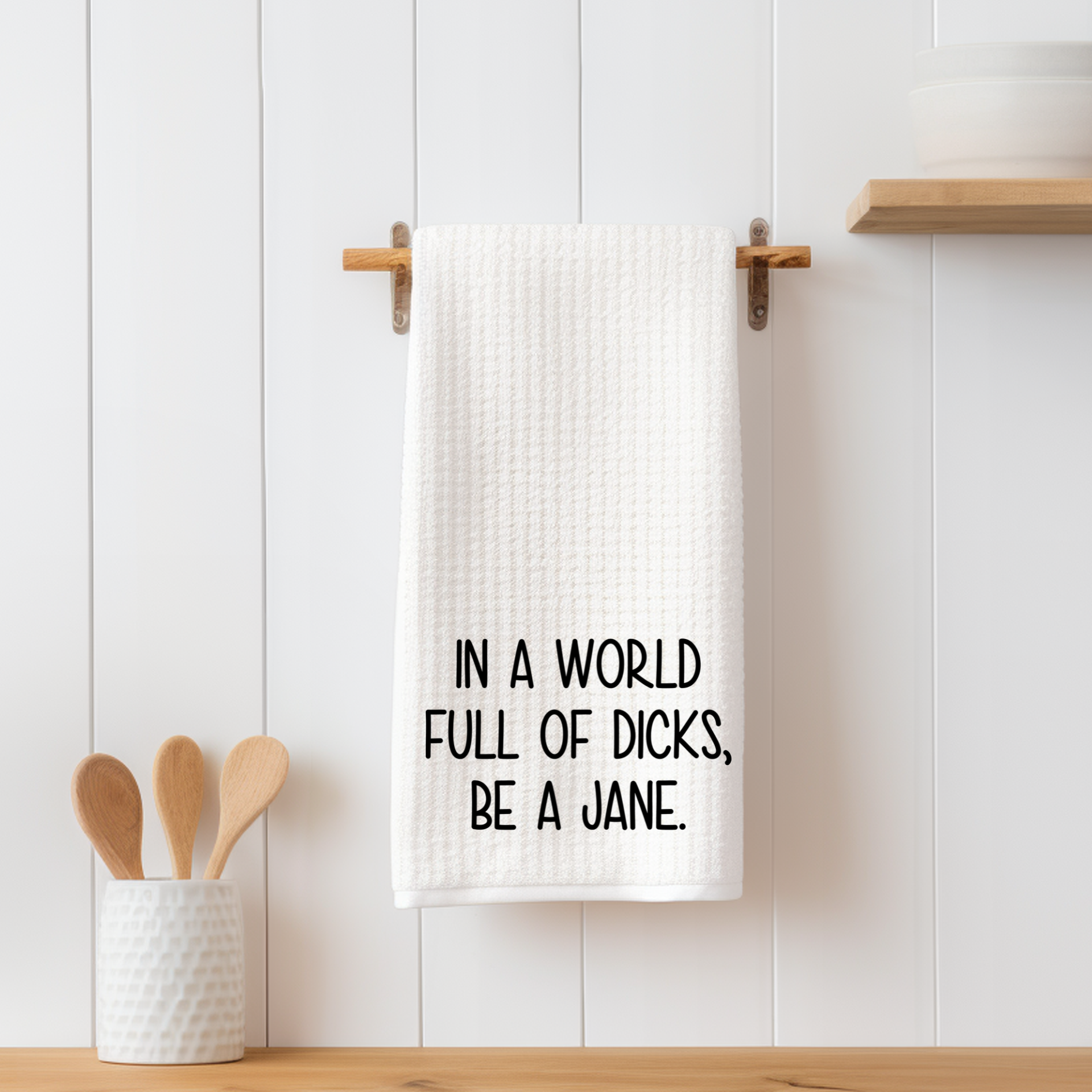 Funny white waffle weave dish towel with bold black text reading "In a world full of dicks, be a Jane," hanging on a wooden peg.