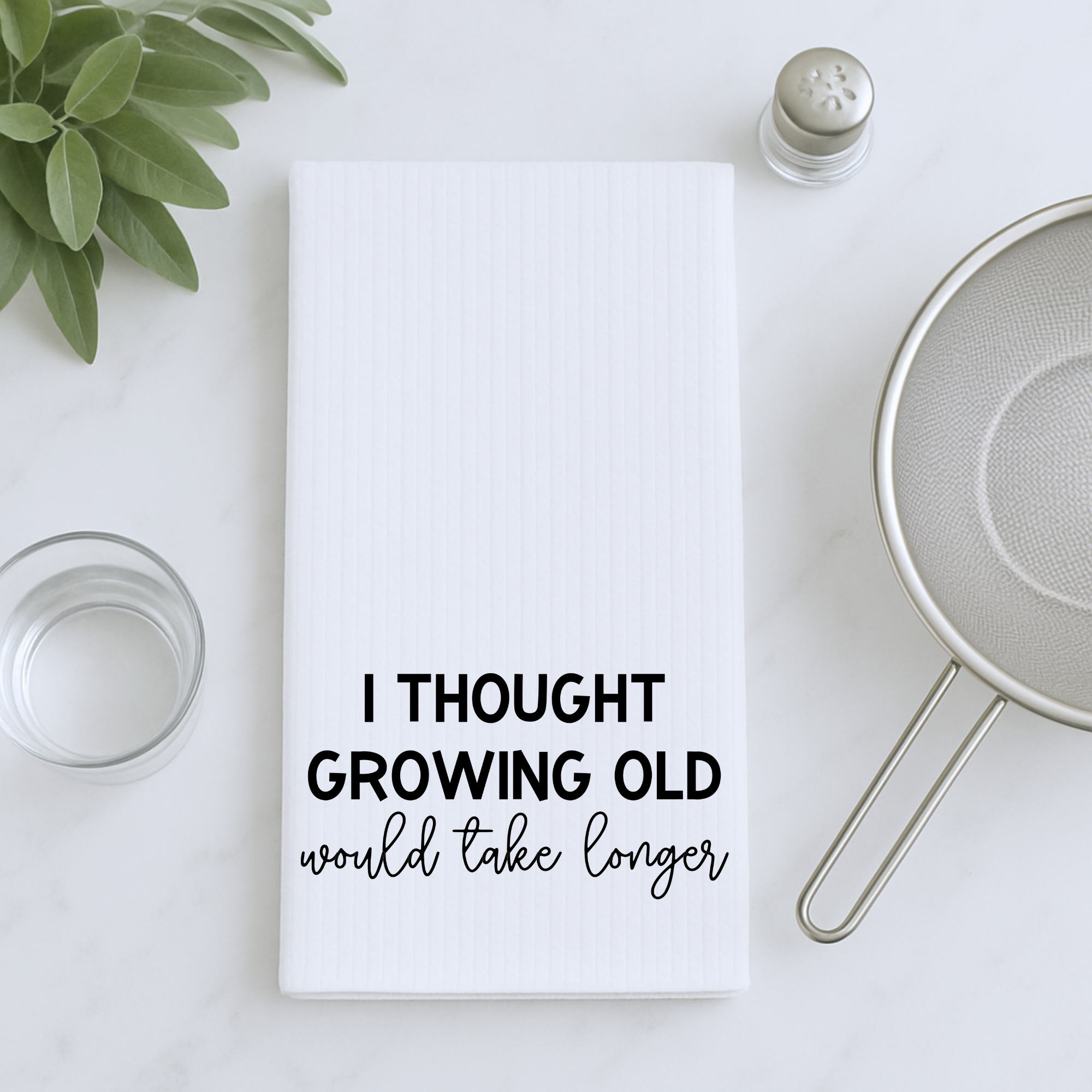 White waffle weave dish towel with funny aging quote styled with herbs and strainer.