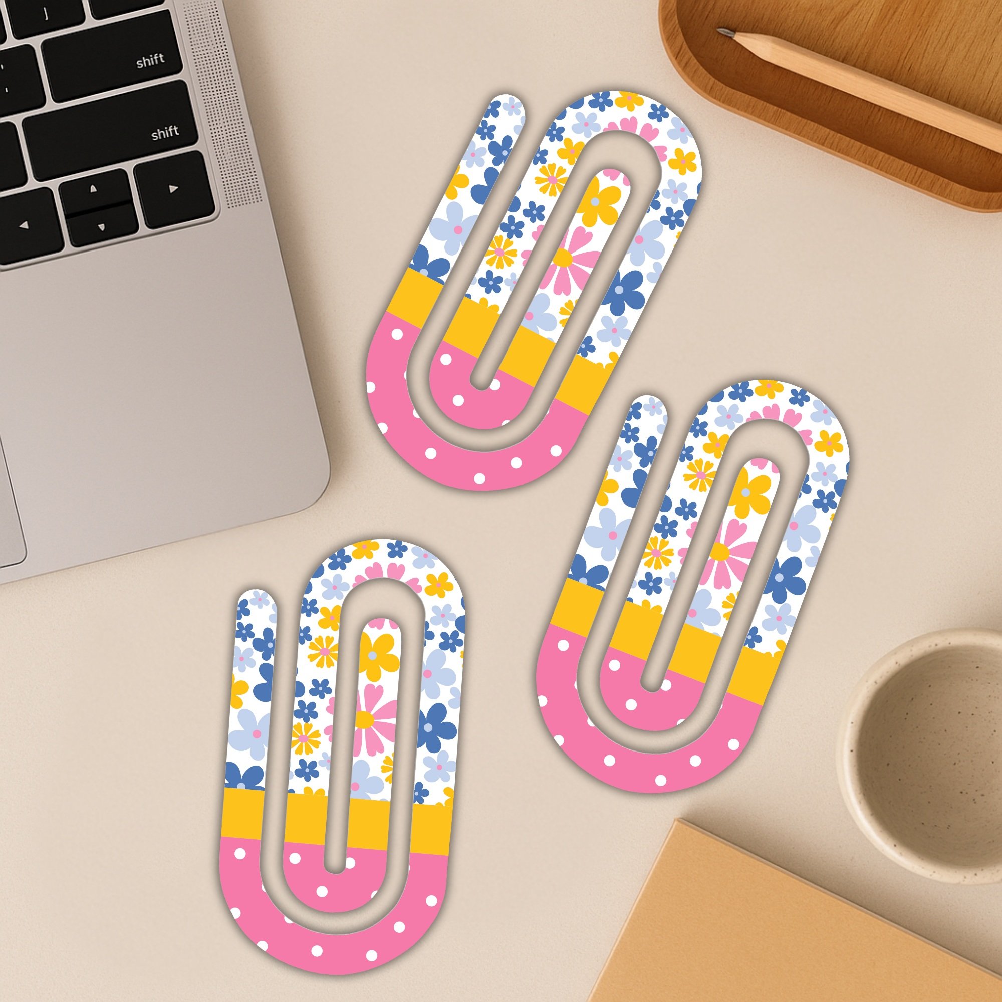 Three colorful jumbo paper clips on a beige desk beside a laptop and coffee cup.