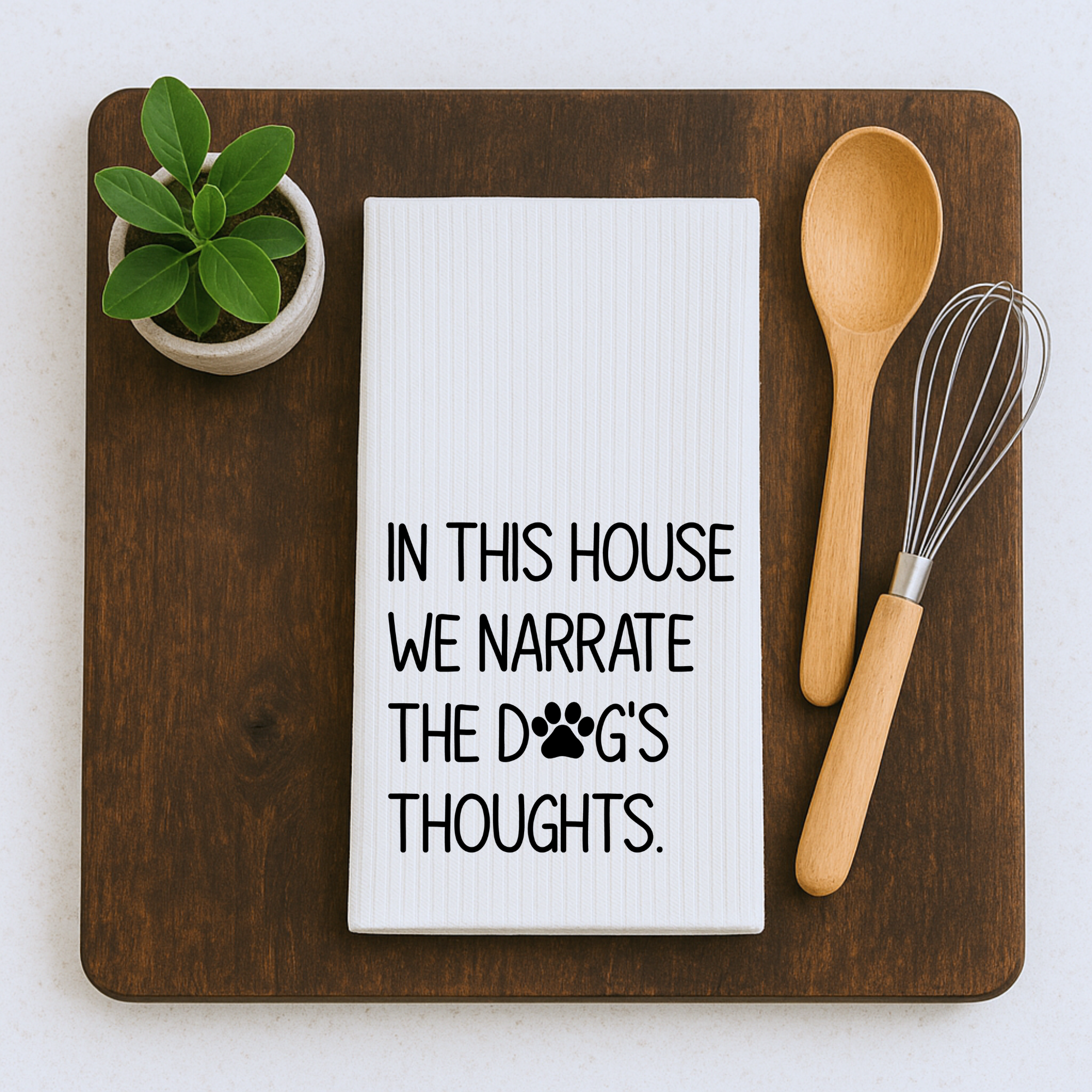 Funny dog quote tea towel on a cutting board styled with whisk, spoon, and small green plant decor.