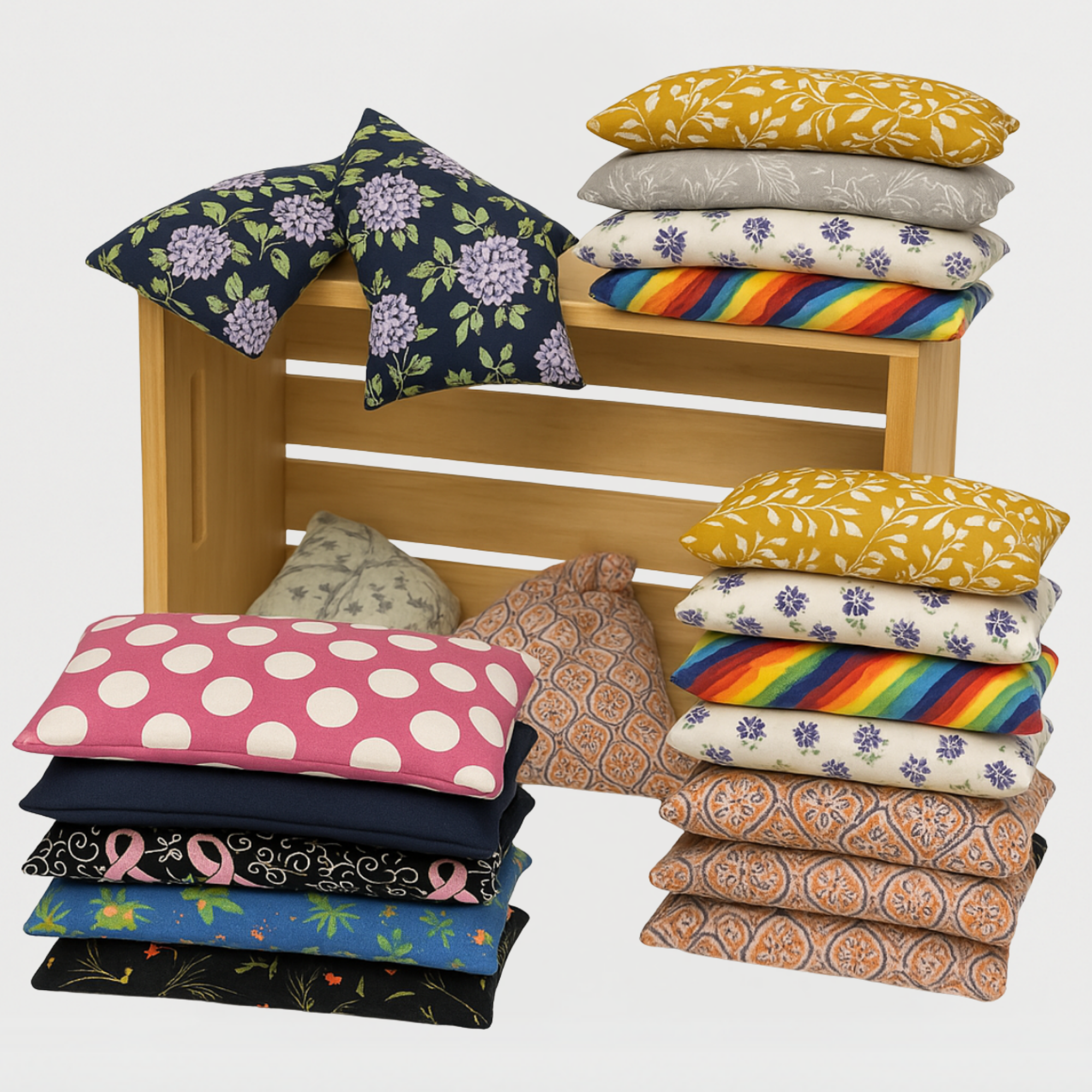 Variety of handmade microwave heat pads with colorful, floral, rainbow, polka dot, and awareness ribbon fabrics stacked beside wooden crate.