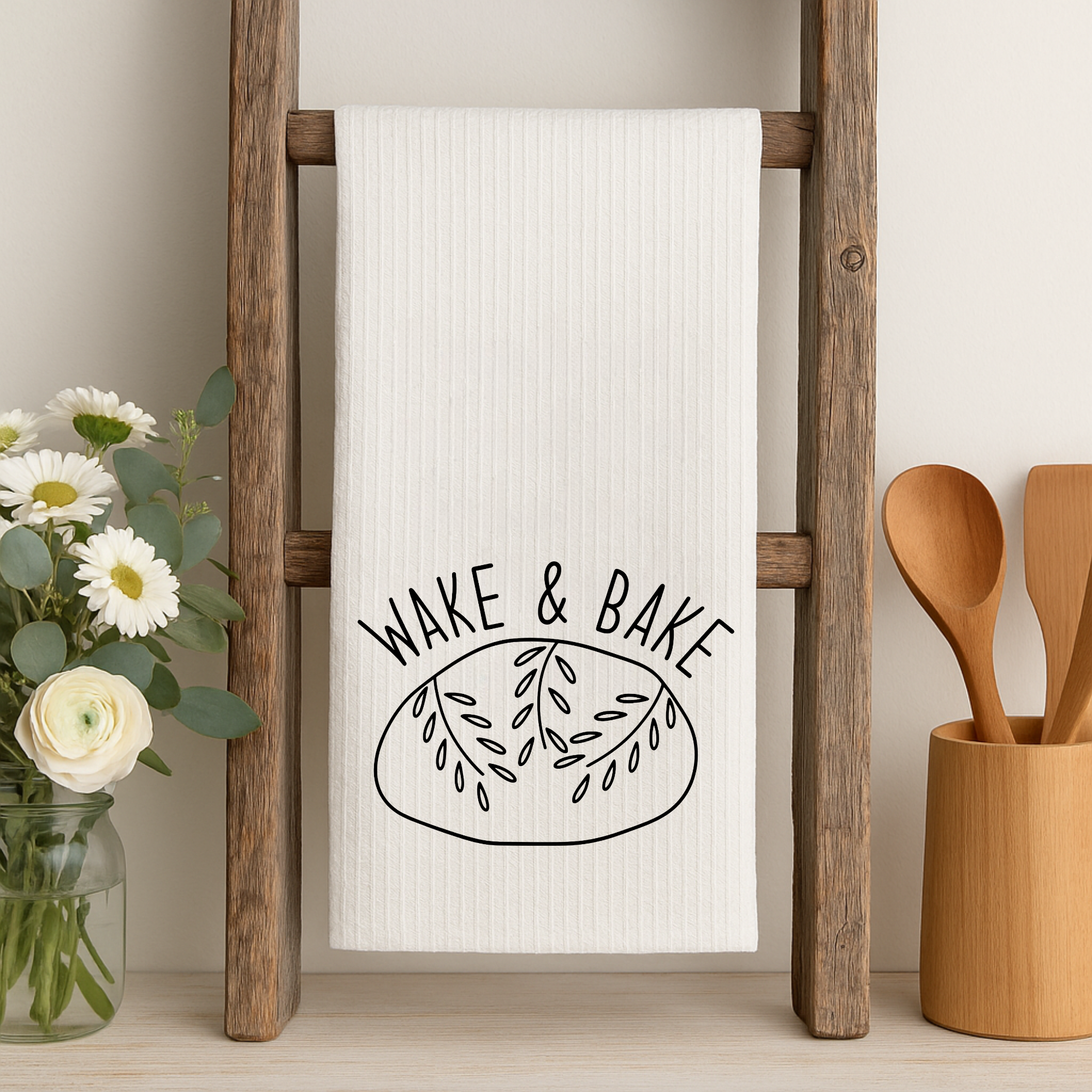 Funny sourdough bread kitchen towel hanging on rustic wood ladder next to flower vase and utensils.