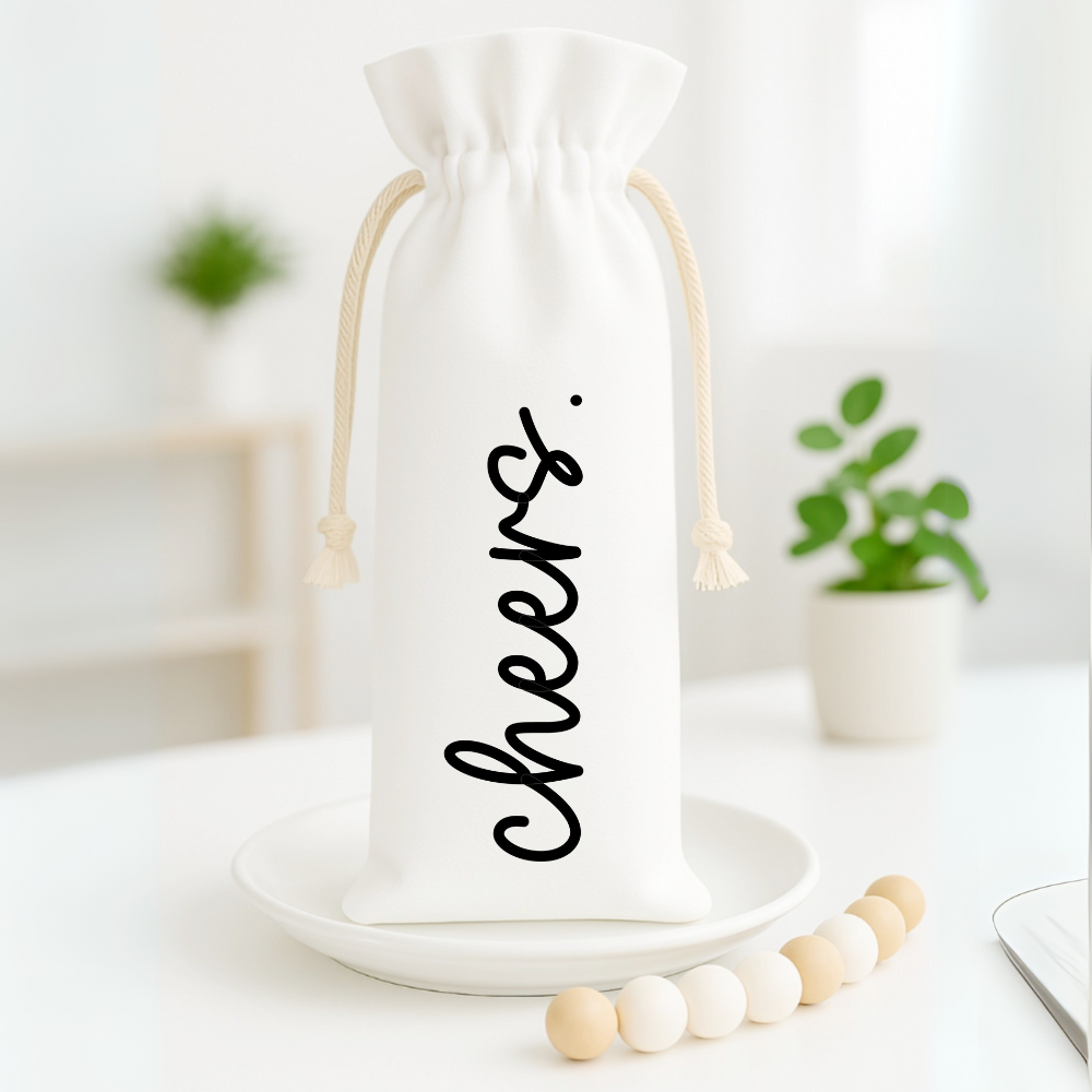 White wine gift bag with “Cheers.” printed in bold black script, displayed on a plate with wooden beads and a plant in soft focus.
