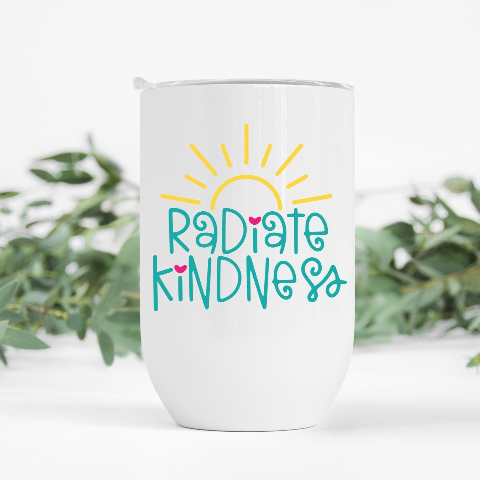 "Radiate Kindness" white wine tumbler with green leaves in the background – Insulated cup for hot and cold drinks, uplifting gift idea.