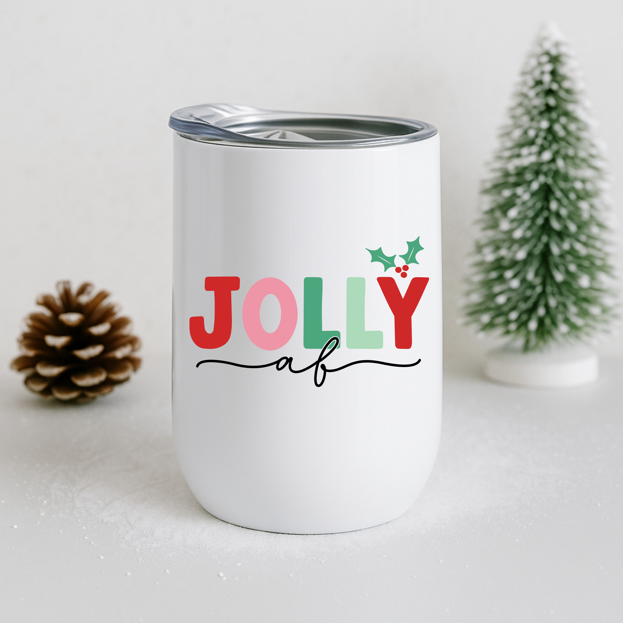 Jolly AF tumbler displayed with a pinecone and frosted tree for a winter aesthetic.