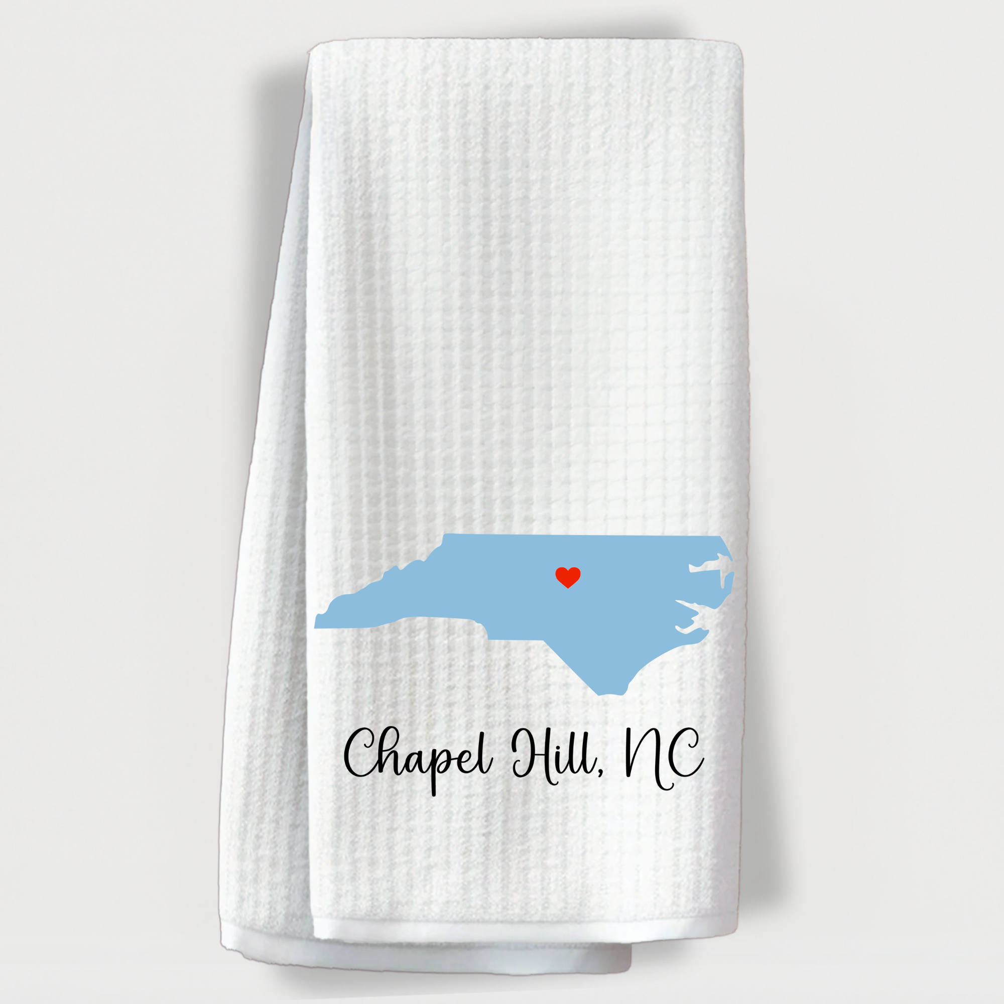 "White towel with blue North Carolina state silhouette and a red heart over Chapel Hill, NC."