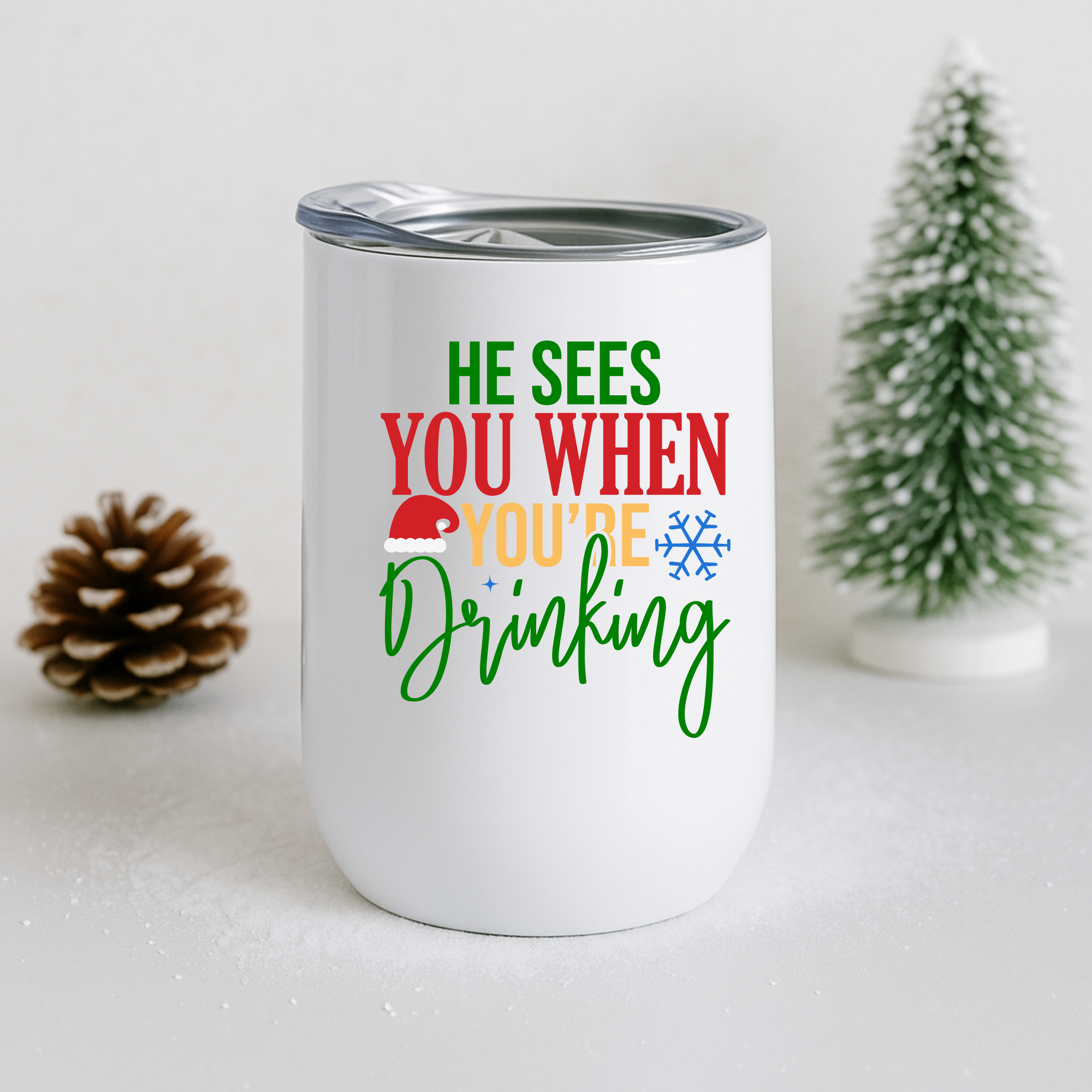 White insulated tumbler with Christmas drinking quote next to pinecone and frosted mini tree.