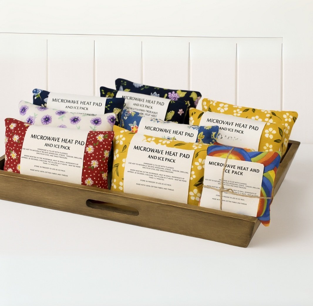 Handmade microwave flaxseed heat pads in colorful floral fabric designs displayed in wooden tray.