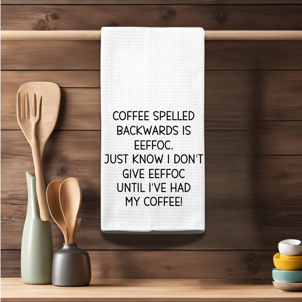 Coffee-themed kitchen towel hanging on a wooden rack, showcasing a humorous quote about coffee spelled backward.