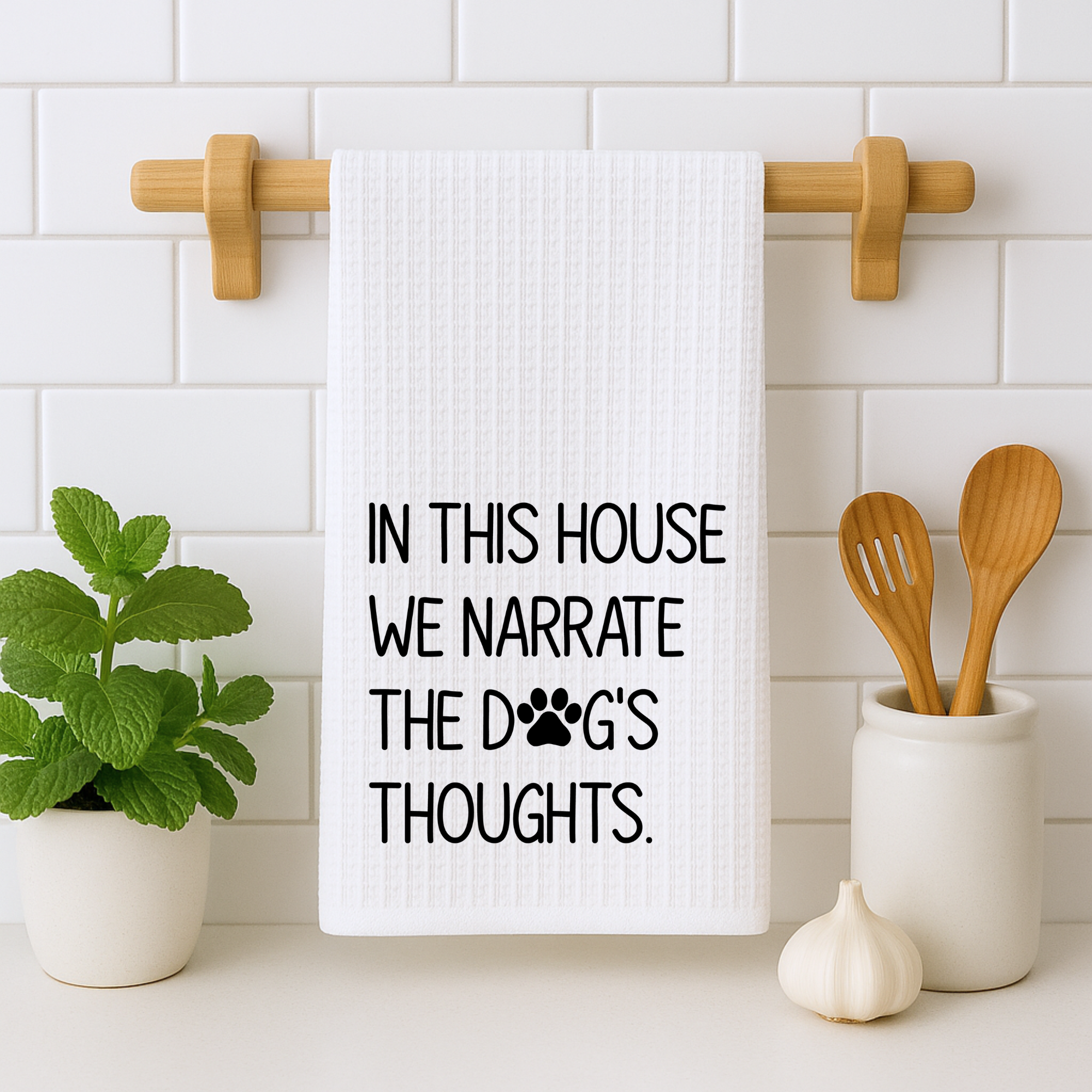 Funny pet humor dish towel displayed on a wooden rack with potted mint and kitchen utensils.