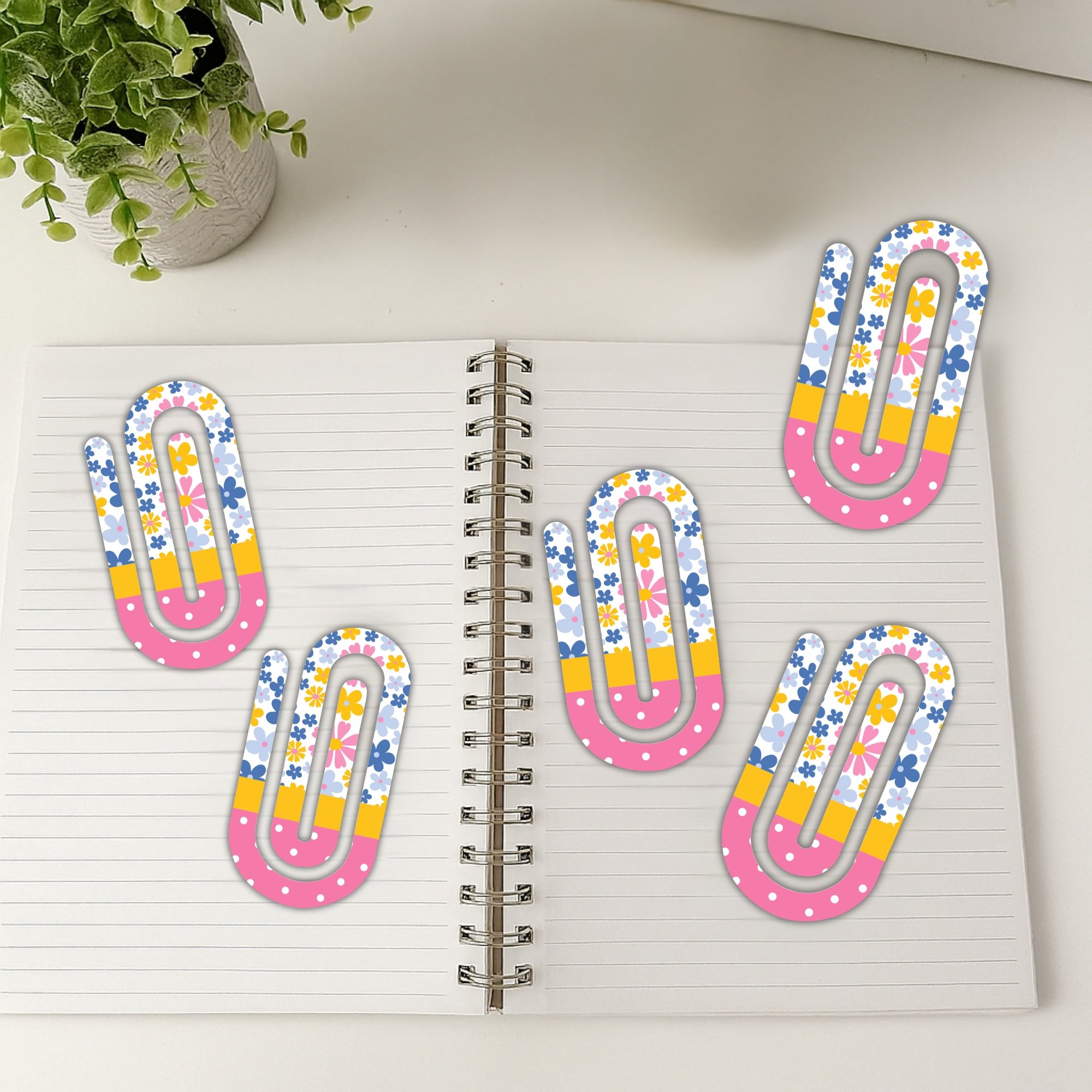 Five large decorative paper clips spread across an open spiral notebook with plant in background.