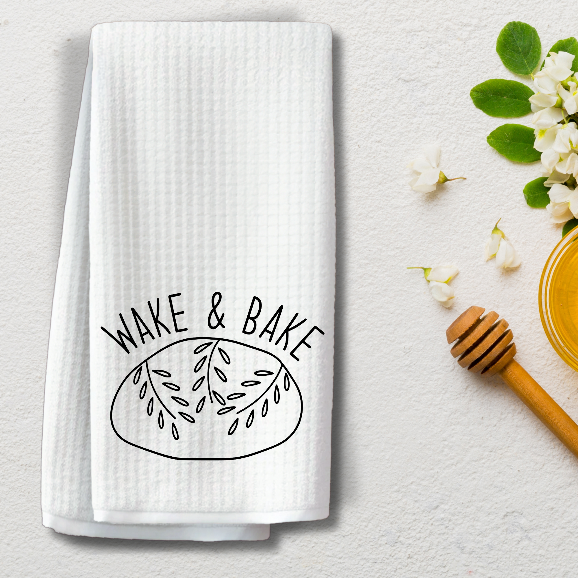 Wake & Bake tea towel styled on textured surface with honey dipper and white blossoms.
