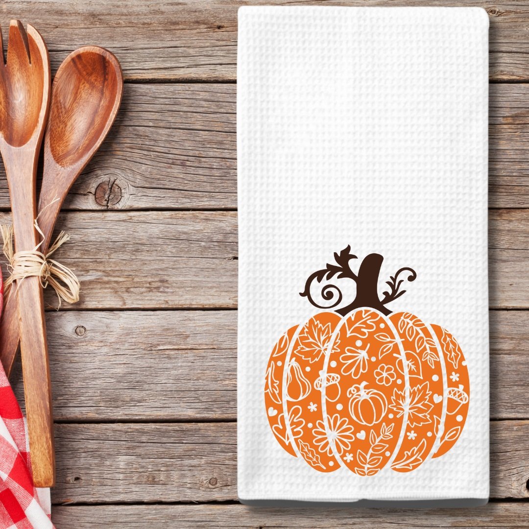 A farmhouse-style autumn kitchen towel featuring a festive orange pumpkin design, displayed on a rustic wooden table alongside wooden spoons tied with twine and a red gingham napkin