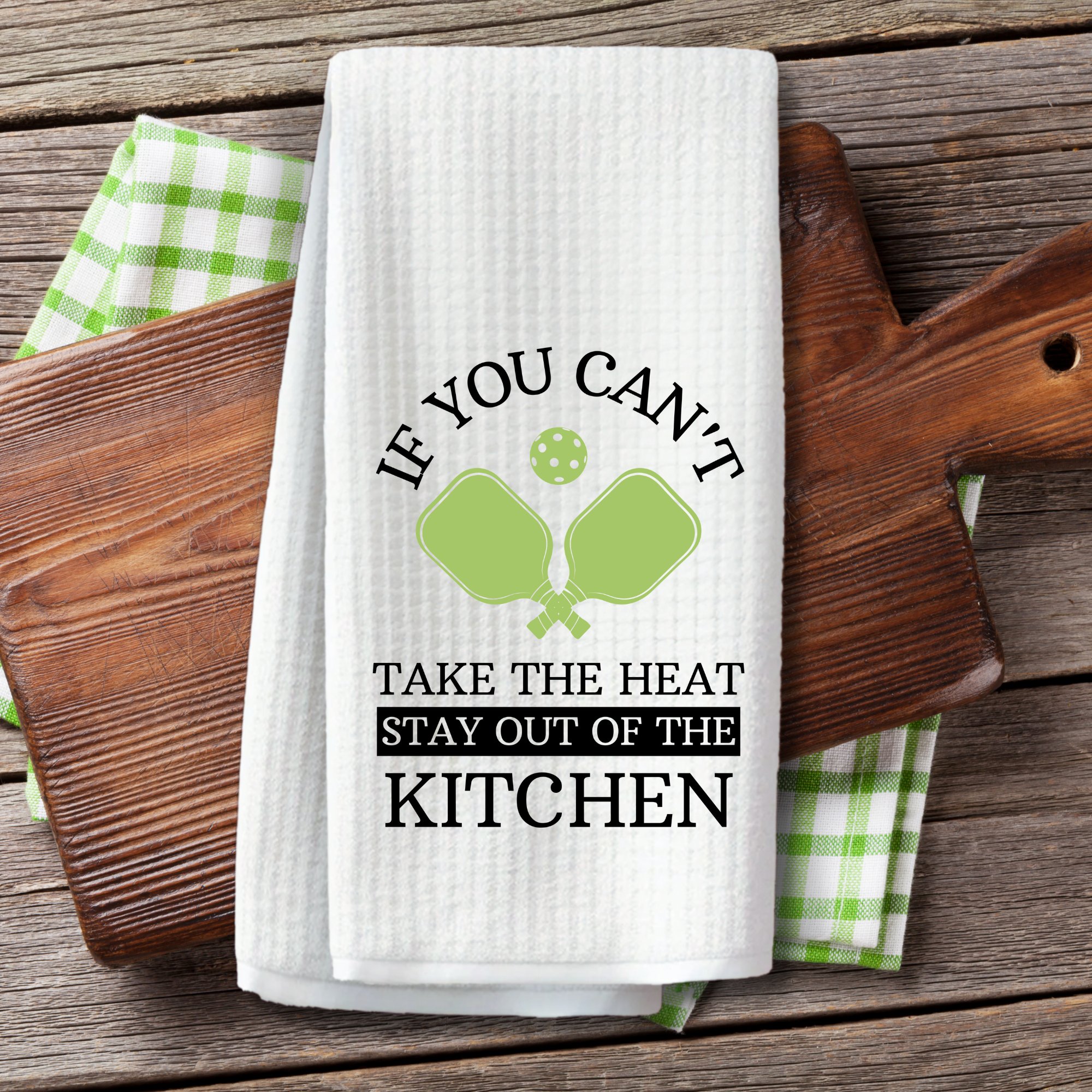 Pickleball-themed dish towel styled with wooden cutting boards and a green gingham towel on rustic wood.