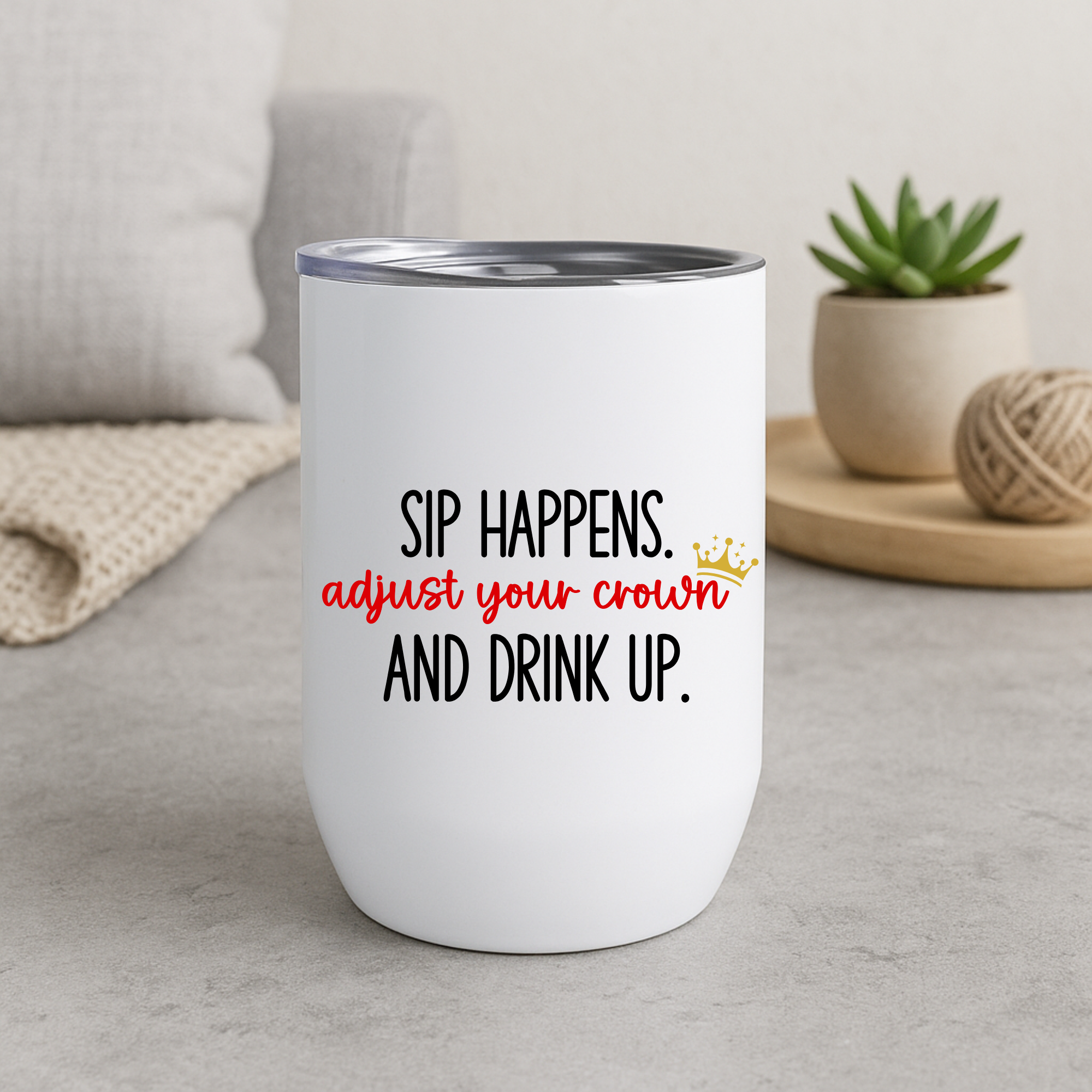 Sip Happens wine tumbler with crown design in red and black text, indoor scene.