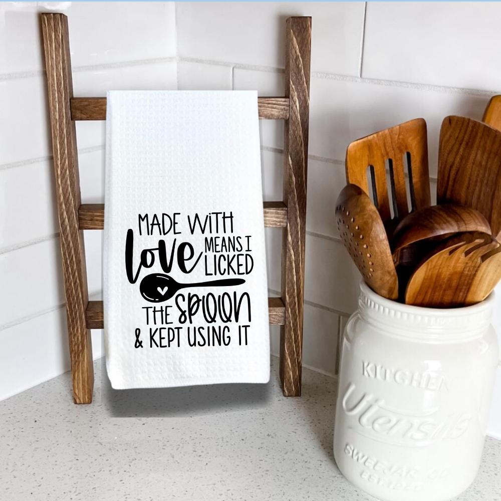 White waffle weave kitchen towel hanging on wooden ladder with funny cooking quote: “Made with love means I licked the spoon and kept using it.”