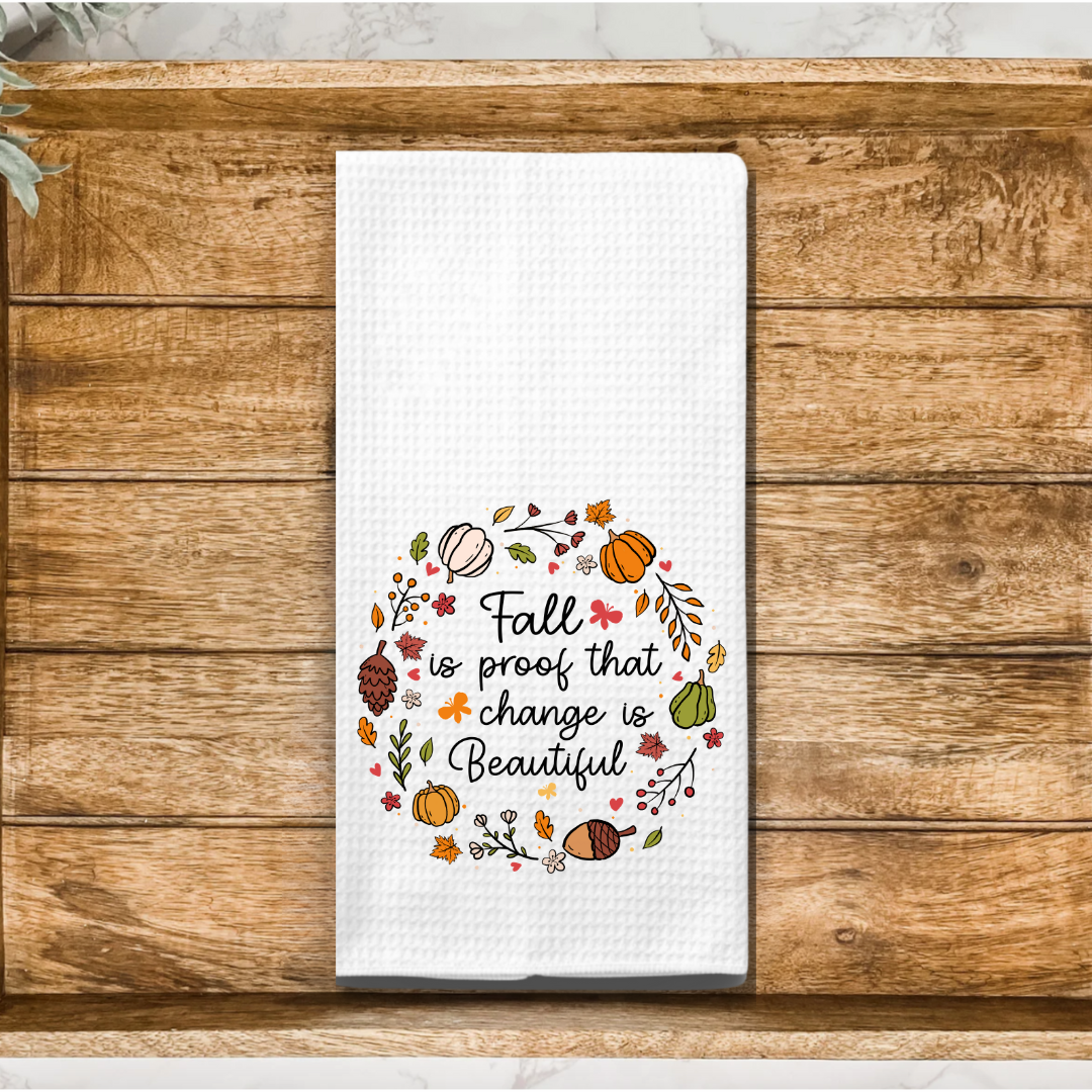 A flat-lay image of a fall-themed kitchen towel with an autumnal wreath design and an inspiring quote, placed on a rustic wooden surface, highlighting the detailed embroidery.