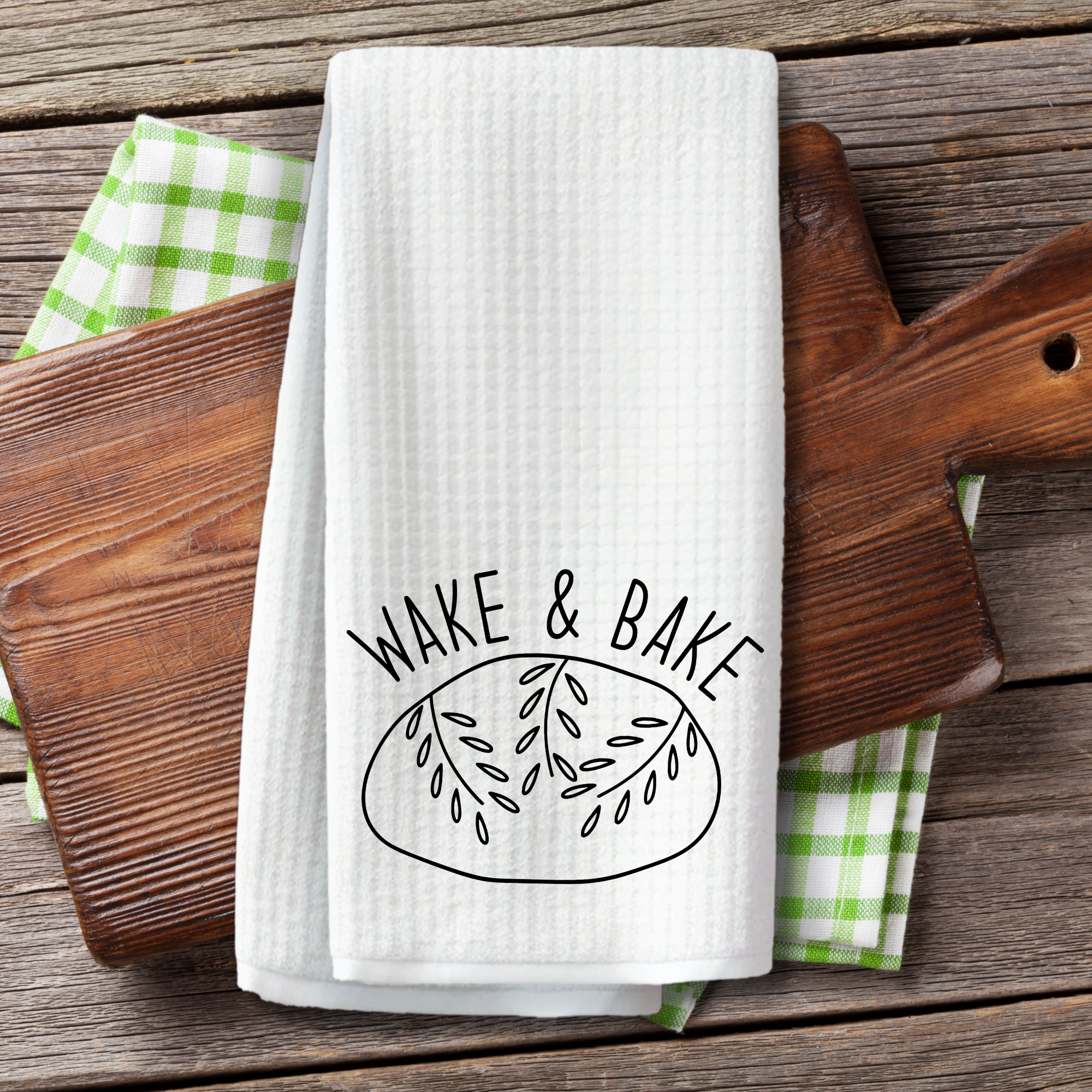 Wake and Bake towel displayed on cutting board and green checkered cloth on rustic wood table.