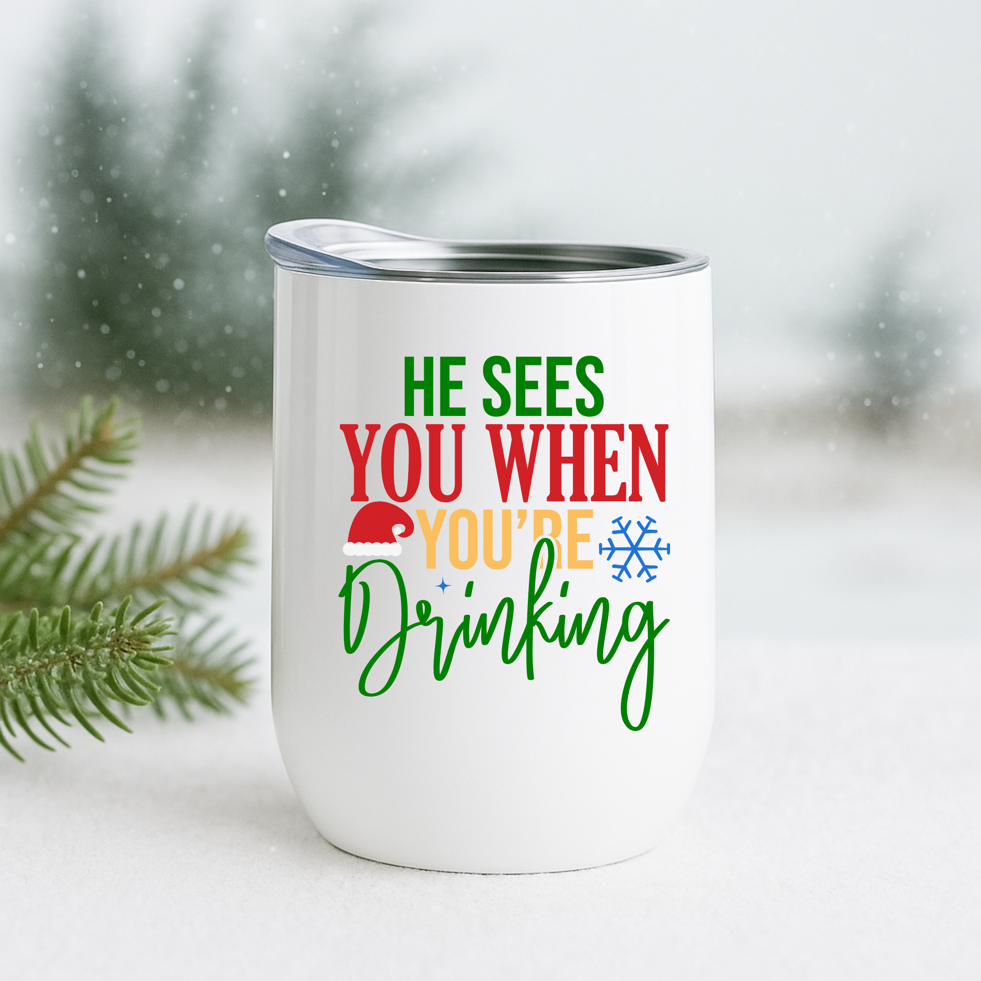 White insulated holiday tumbler with the phrase “He Sees You When You’re Drinking,” displayed in a snowy winter scene with pine branches.