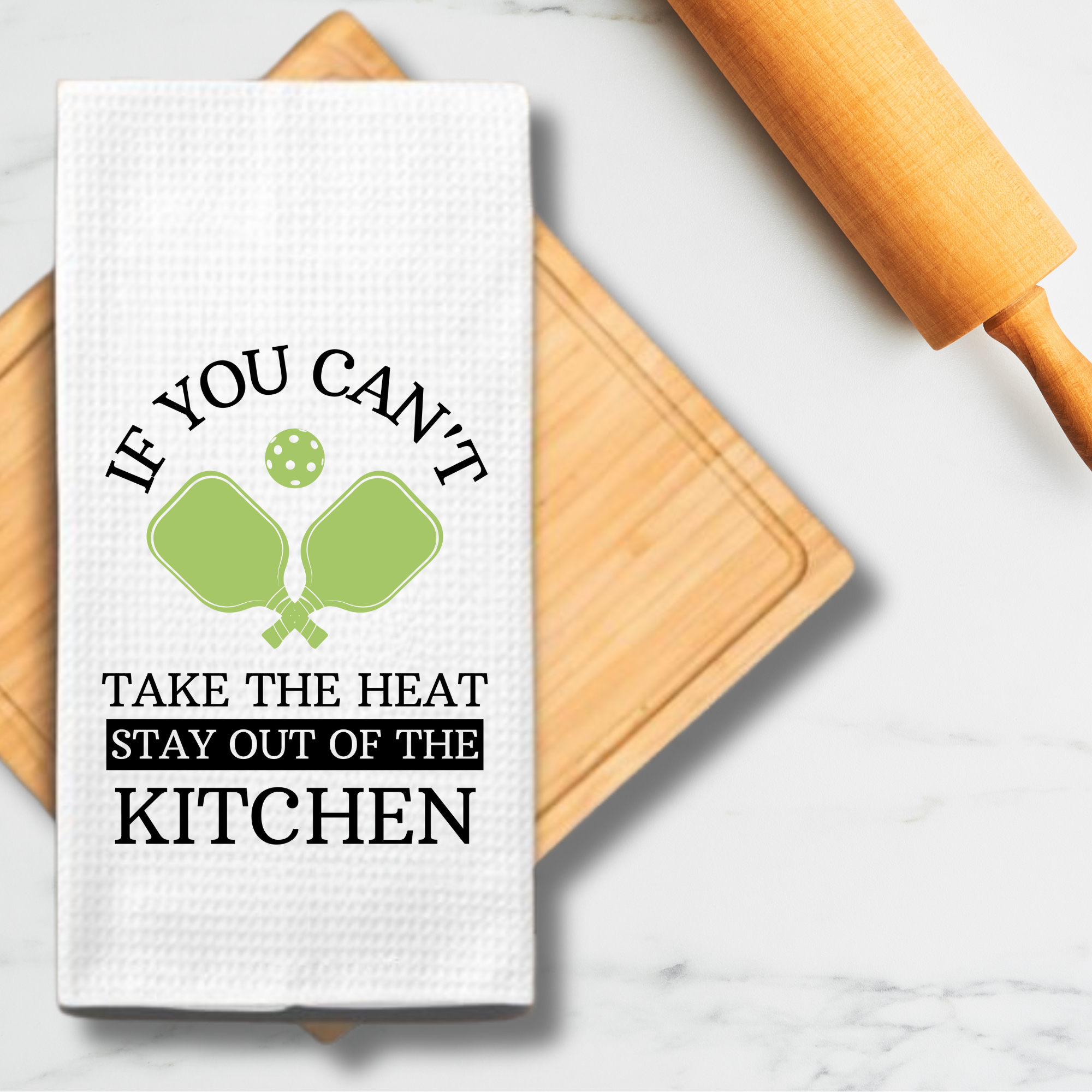 Kitchen towel with green pickleball paddles displayed on a cutting board with rolling pin and marble counter.