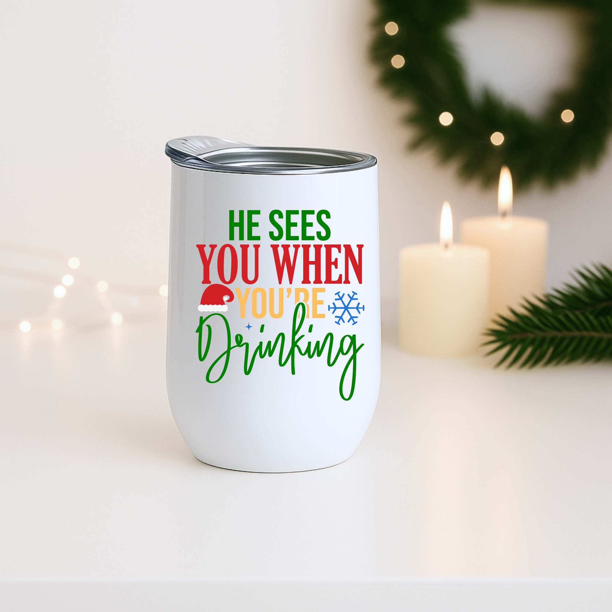 White tumbler with holiday drinking quote displayed beside candles and greenery with warm Christmas lighting.