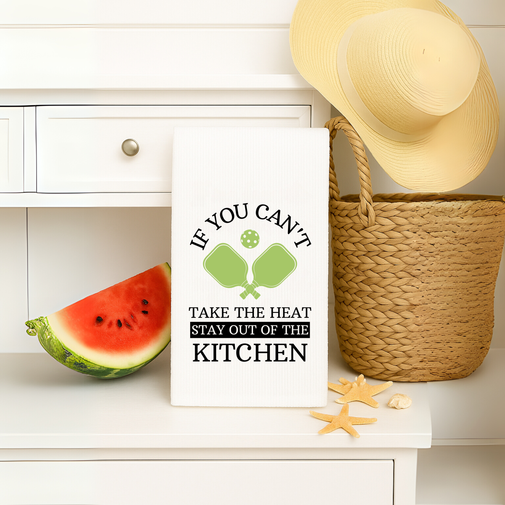 Funny pickleball-themed towel on a white cabinet with a straw hat, watermelon, and beach decor.