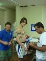 A woman in hospital gown sitting on a bed, with two men, one in blue scrubs and one in white, attending to her in a hospital room.