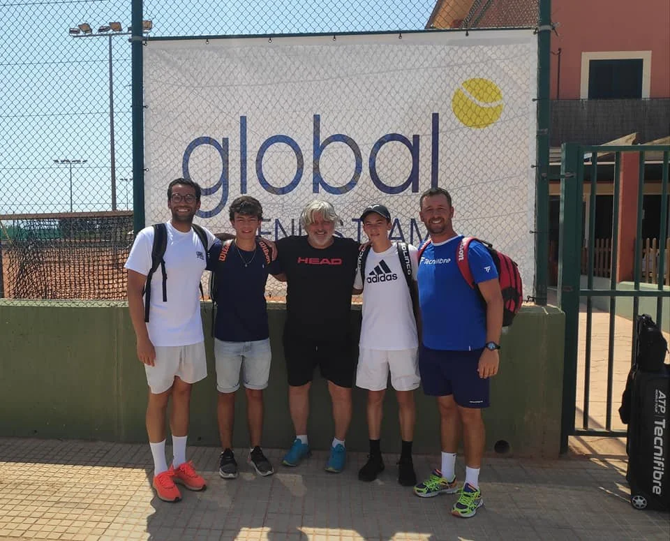 Group of five people standing in front of a tennis court fence and a large sign that says 'global' on a sunny day.