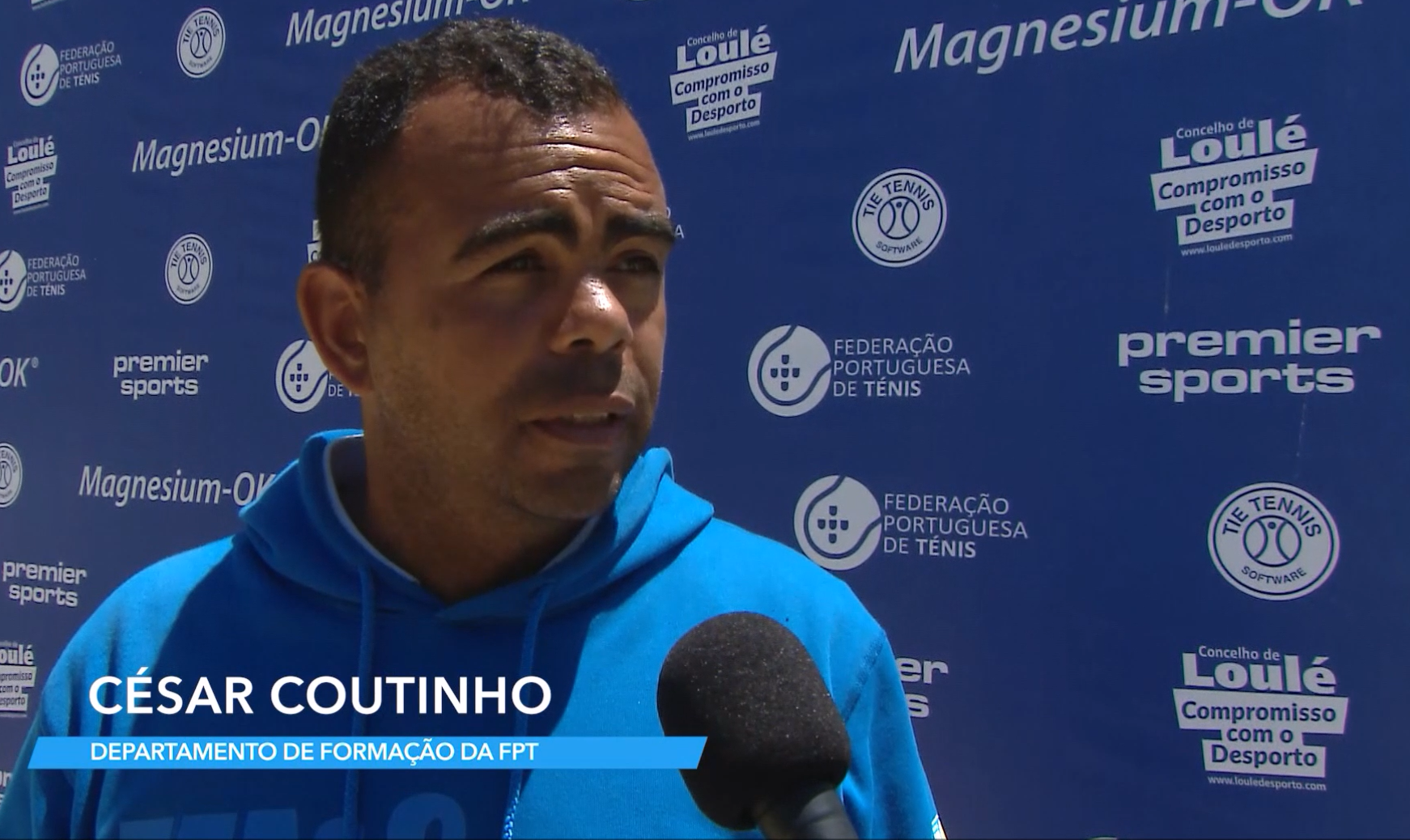 César Coutinho being interviewed outdoors with a blue backdrop featuring logos from Portuguese tennis organizations and sponsors