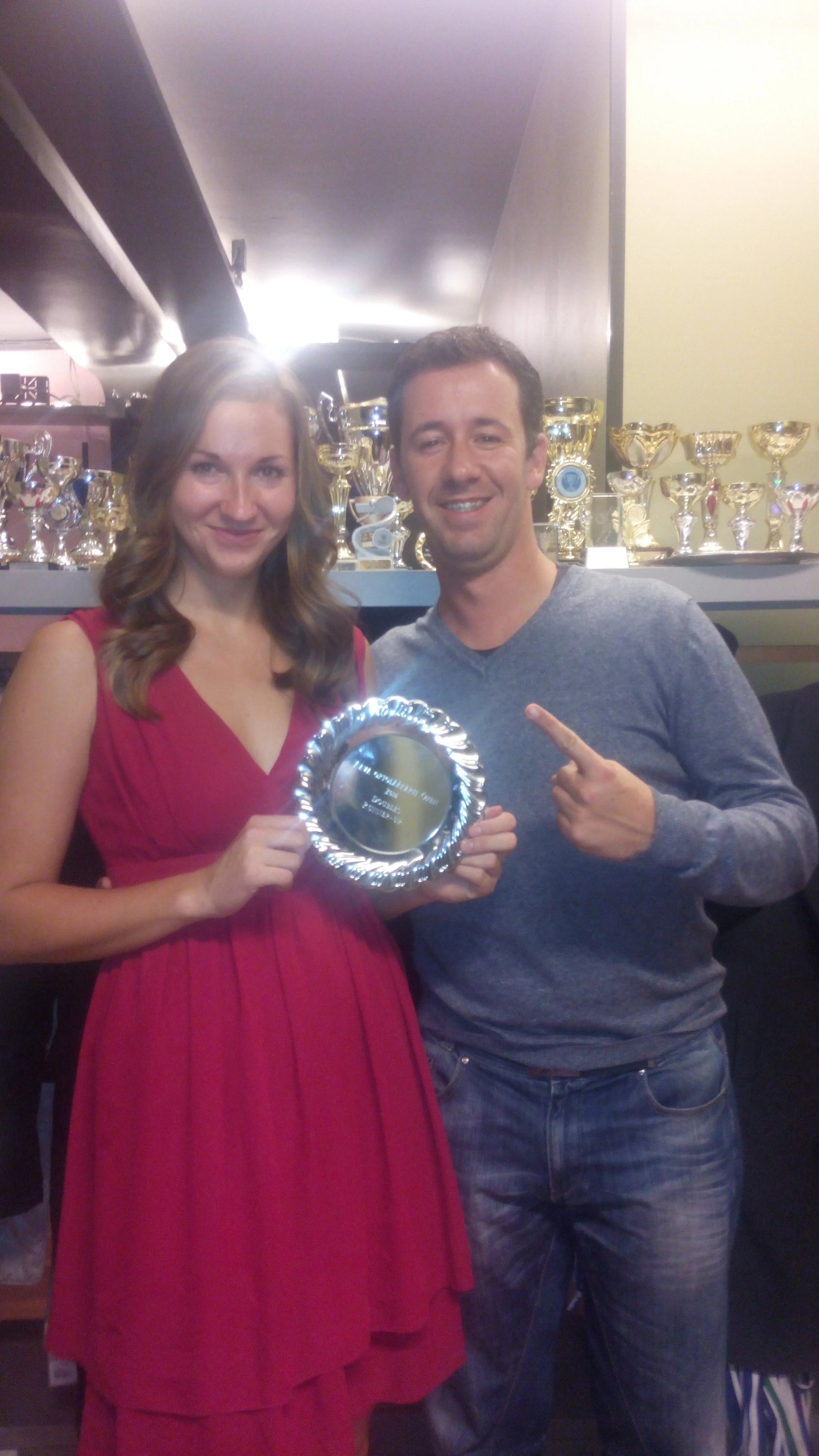 A woman in a red dress and a man in a gray shirt are smiling and holding a shiny silver plate, standing in front of a shelf filled with numerous trophies.