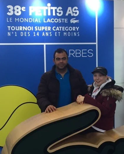 A man and a girl smiling and posing at a golf event, with a blue backdrop displaying text in French and details about a golf tournament at Le Golf Lacoste.