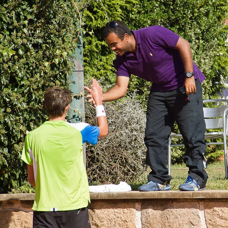 A man in a purple polo shirt and black pants giving a handshake or high five to a young boy wearing a bright green sports jersey outside in front of green bushes.