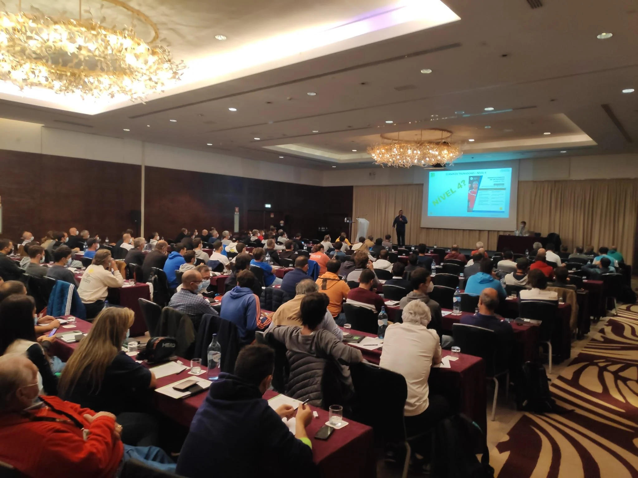 A large conference room filled with many attendees seated at long tables, listening to a speaker at a podium in front of a large projector screen.