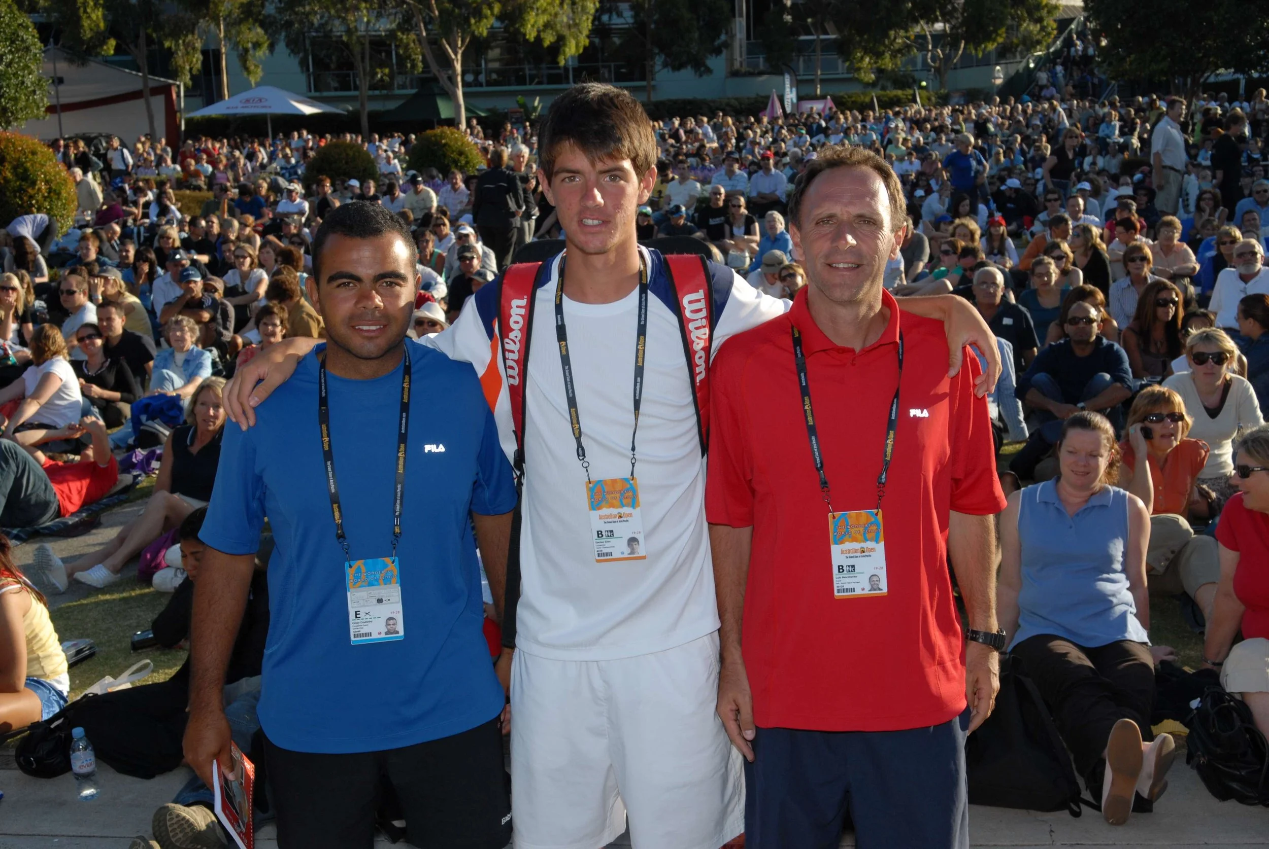 Three men standing outdoors in front of a large seated crowd during a public event, with all wearing casual athletic clothing and event identification badges.
