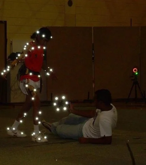 A person dressed with LED lights standing above a person lying on the floor in a dark room with a wooden wall background. There is a camera on a tripod in the background.