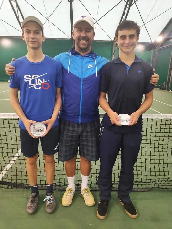 Two young male tennis players holding awards stand with their coach on an indoor tennis court, smiling for a photo.