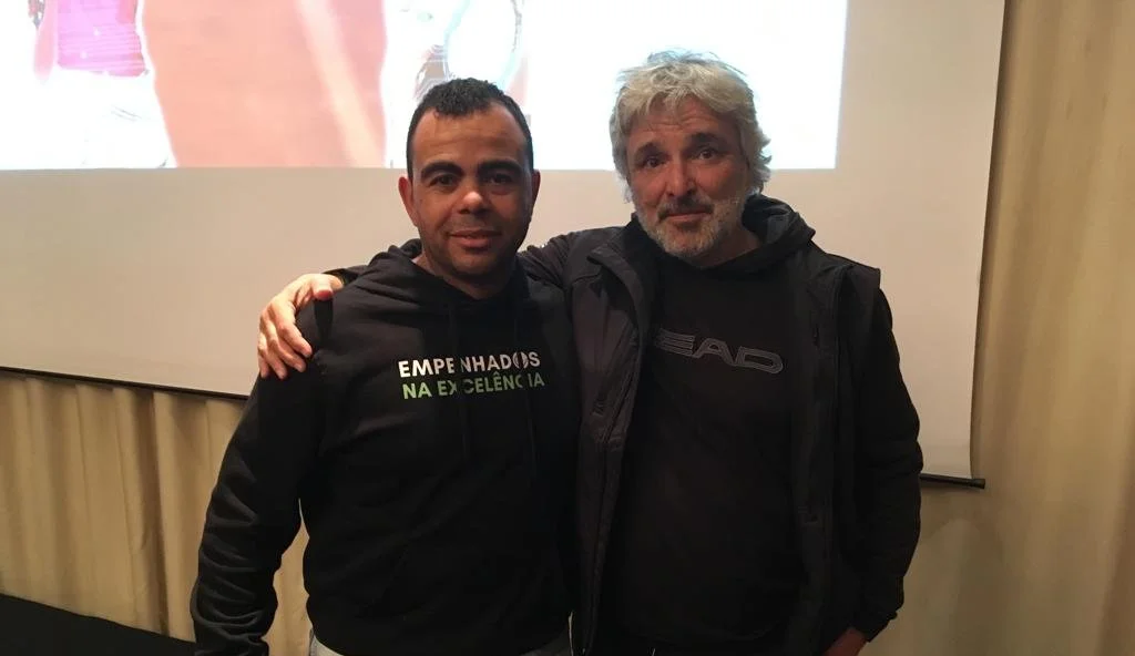 Two men standing close together, both wearing black hoodies, with their arms around each other's shoulders, indoors in front of a projected image.