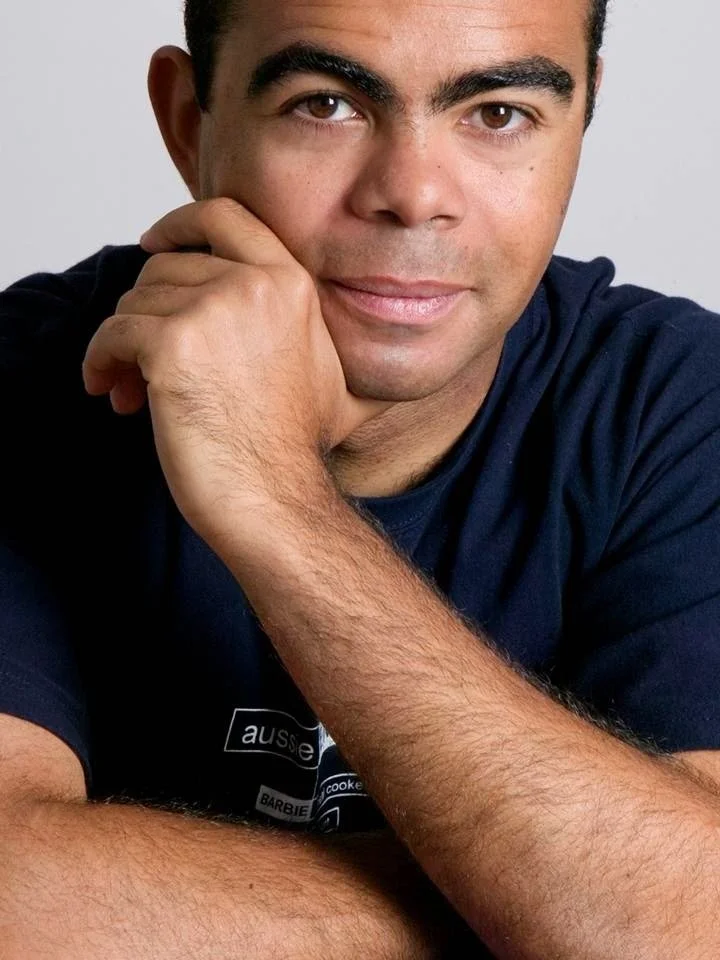 Close-up portrait of a man with short dark hair and brown eyes, resting his chin on his hand, wearing a dark blue T-shirt.