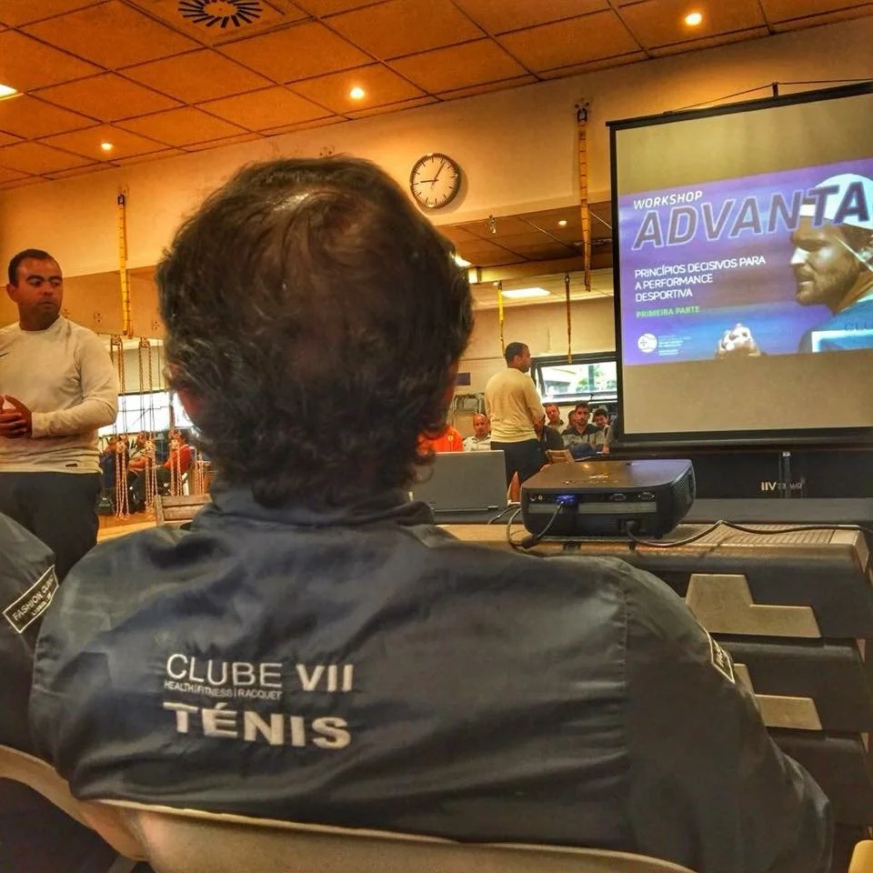 A man with dark hair and wearing a black jacket with white text that says 'CLUBE VII TENIS' is attending a workshop or seminar in a room with multiple people; a presentation slide on the screen shows a sports-related workshop titled 'ADVANTA PRINCIPIOS DECISIVOS PARA A PERFORMANCE DESPORTIVA.'
