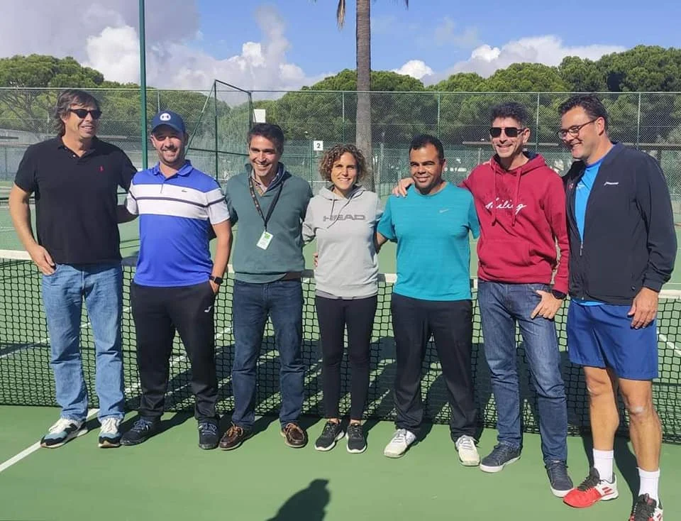 Group of eight people standing on a tennis court, smiling, with a net and green fencing in the background, and trees and blue sky overhead.
